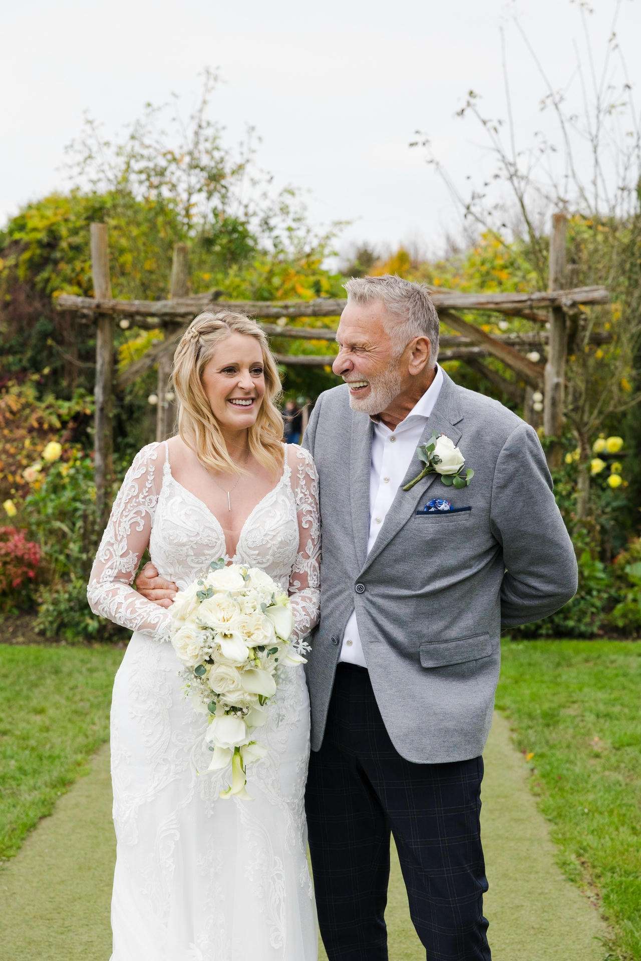 A bride in a white lace wedding dress holds a white bouquet and smiles as she stands with a man in a gray blazer in a garden.