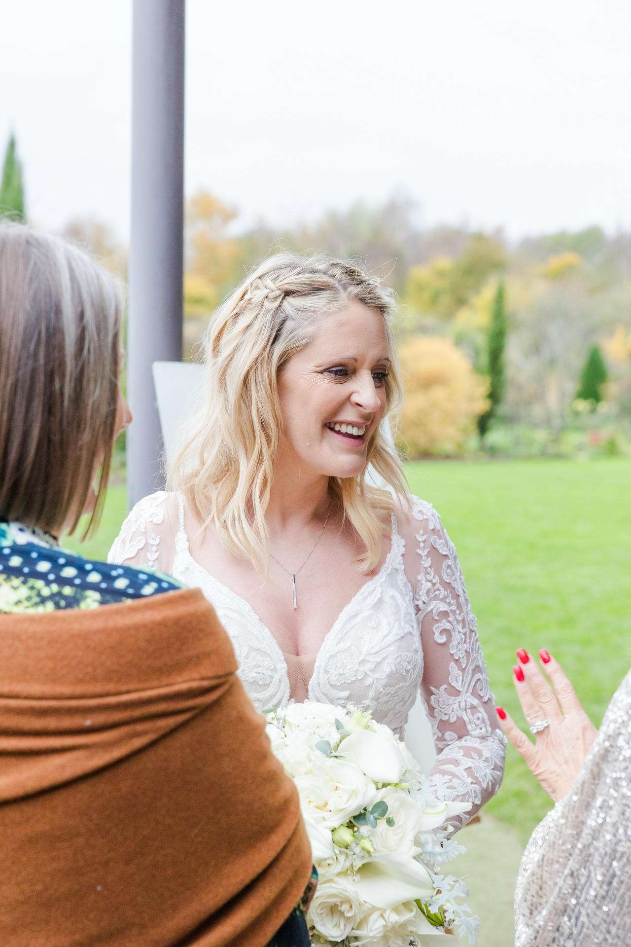 Bride in a lace white wedding gown holding a bouquet of white roses, smiling as she talks with guests outdoors.