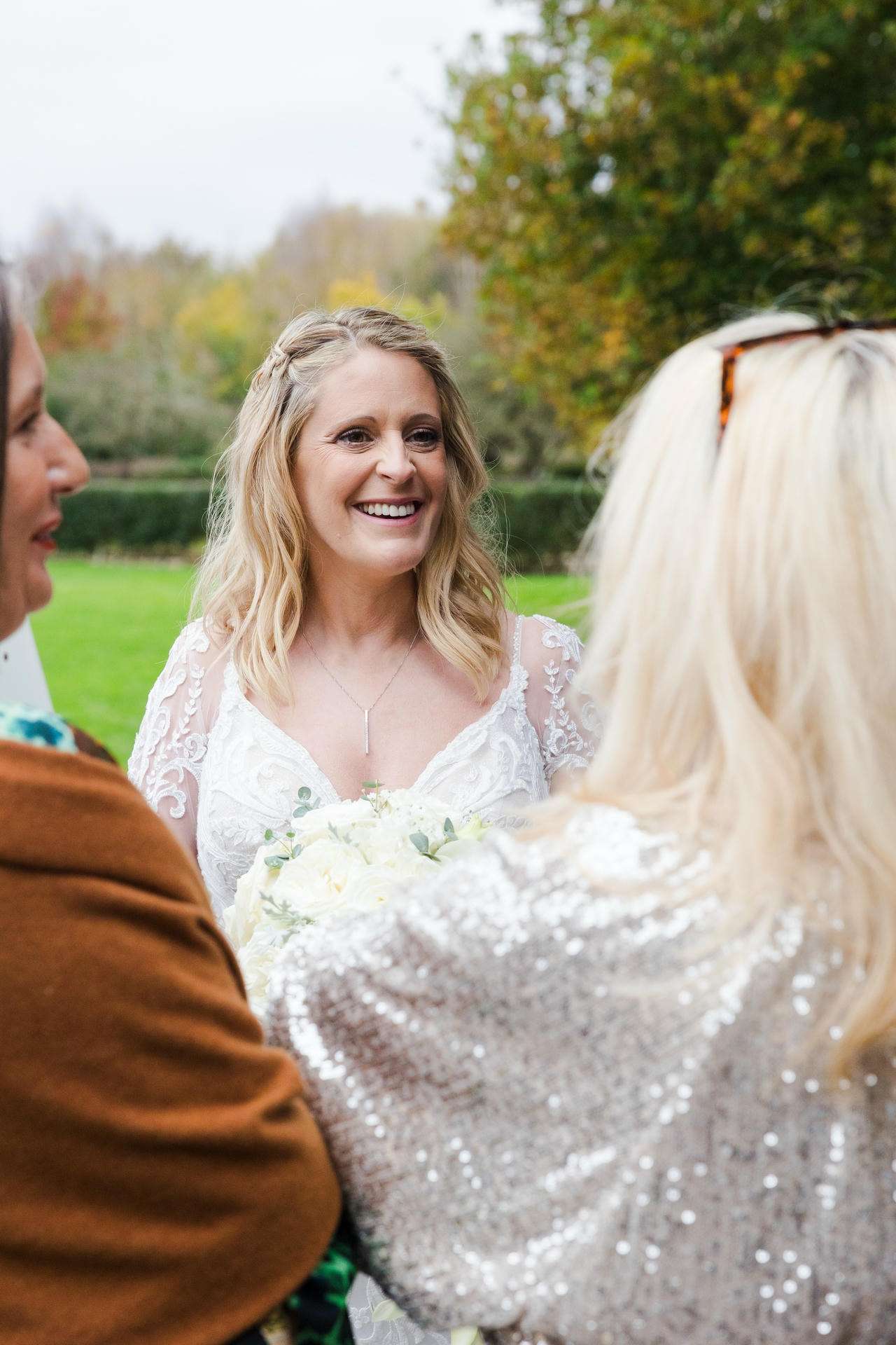 Smiling bride in a lace wedding dress holds a white bouquet while chatting with two guests outdoors.