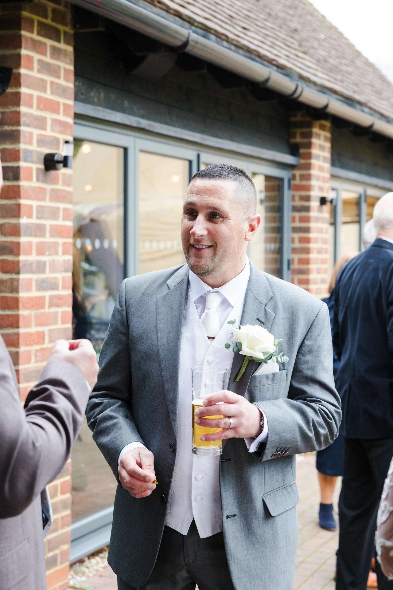 Man in a gray suit with a white rose boutonniere holds a beer and chats outdoors beside a brick building.