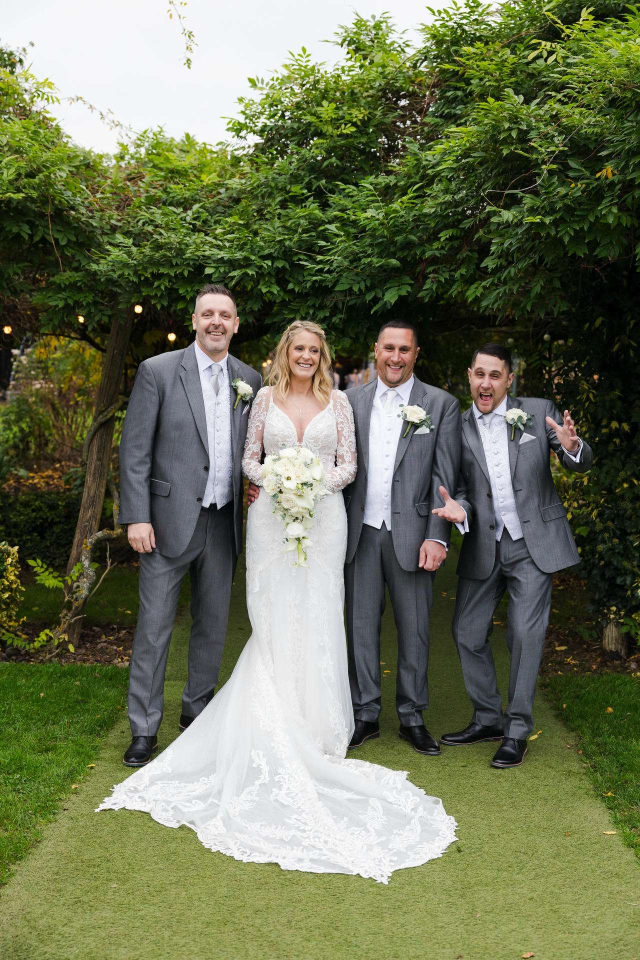 Bride in a white lace wedding gown with a long train poses with three groomsmen in gray suits outdoors among lush green trees.
