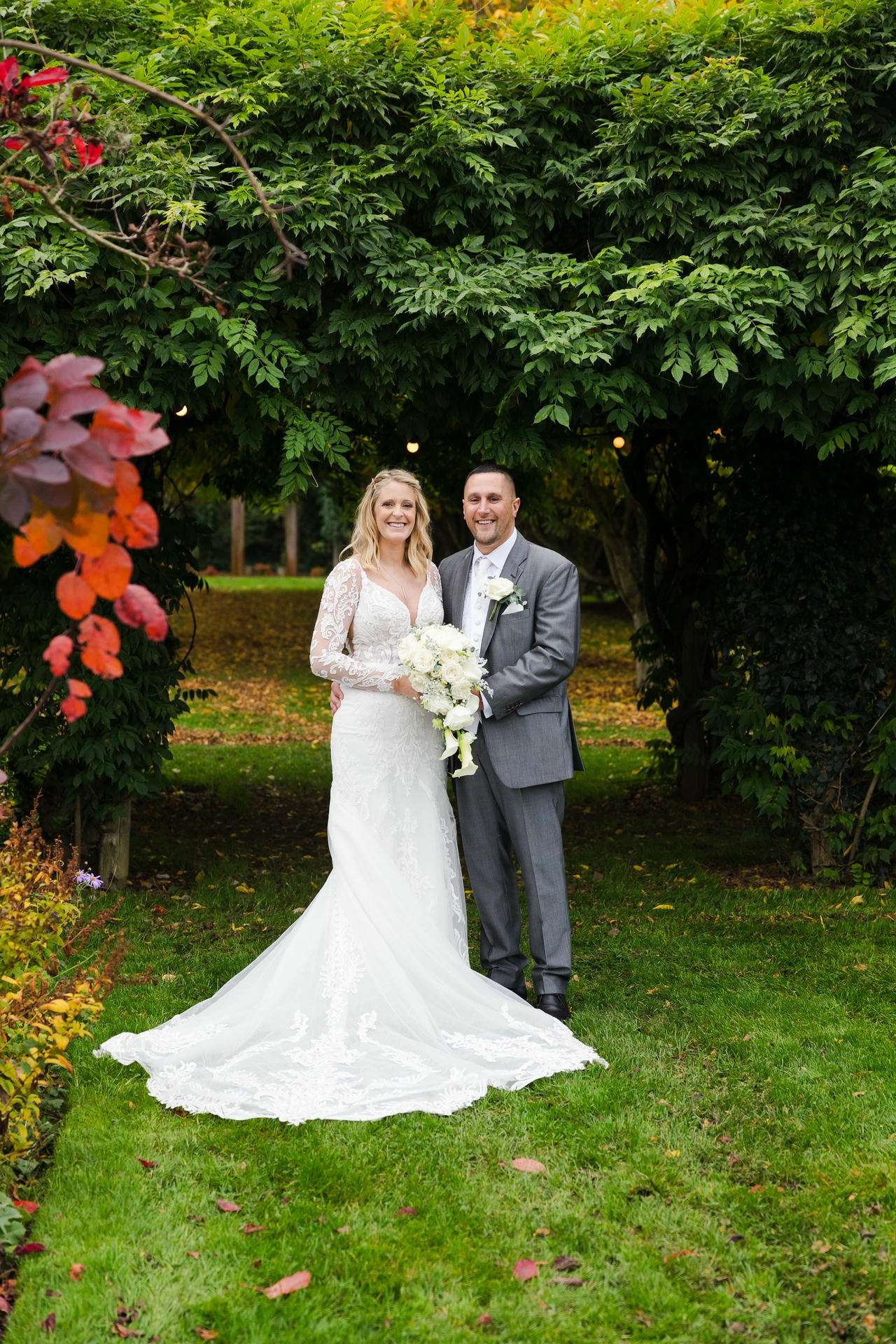 Bride in a white lace gown with long sleeves and a train stands with her groom in a gray suit under a leafy garden arch, holding a white bouquet.