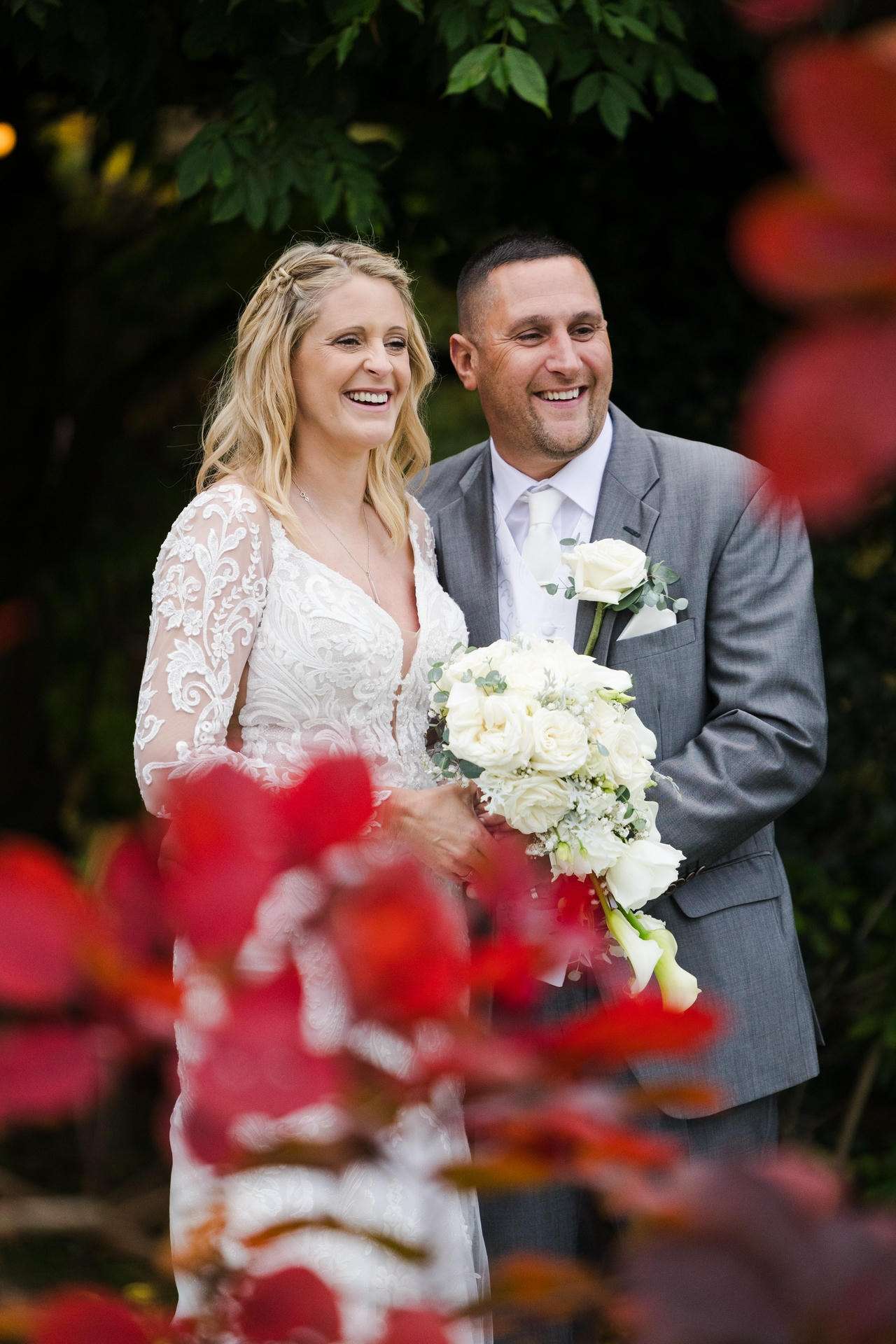 Smiling bride in a lace wedding dress holds a bouquet of white roses beside a groom in a gray suit outdoors at The Gardens Wedding venue