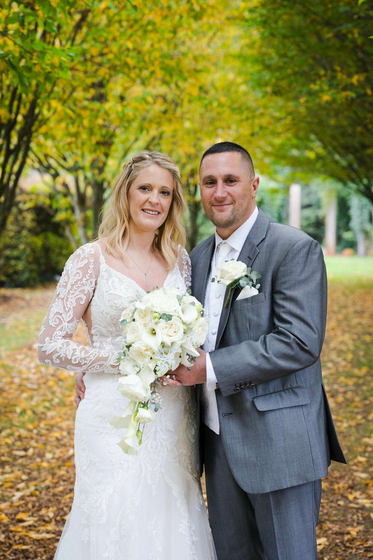 A bride in a white lace wedding dress holding a white bouquet stands with a groom in a gray suit outdoors among autumn leaves.