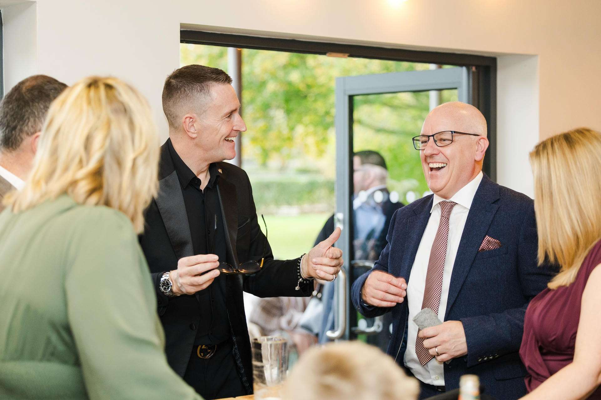 Two men in suits laugh and chat at a social gathering indoors near an open door with greenery visible outside.