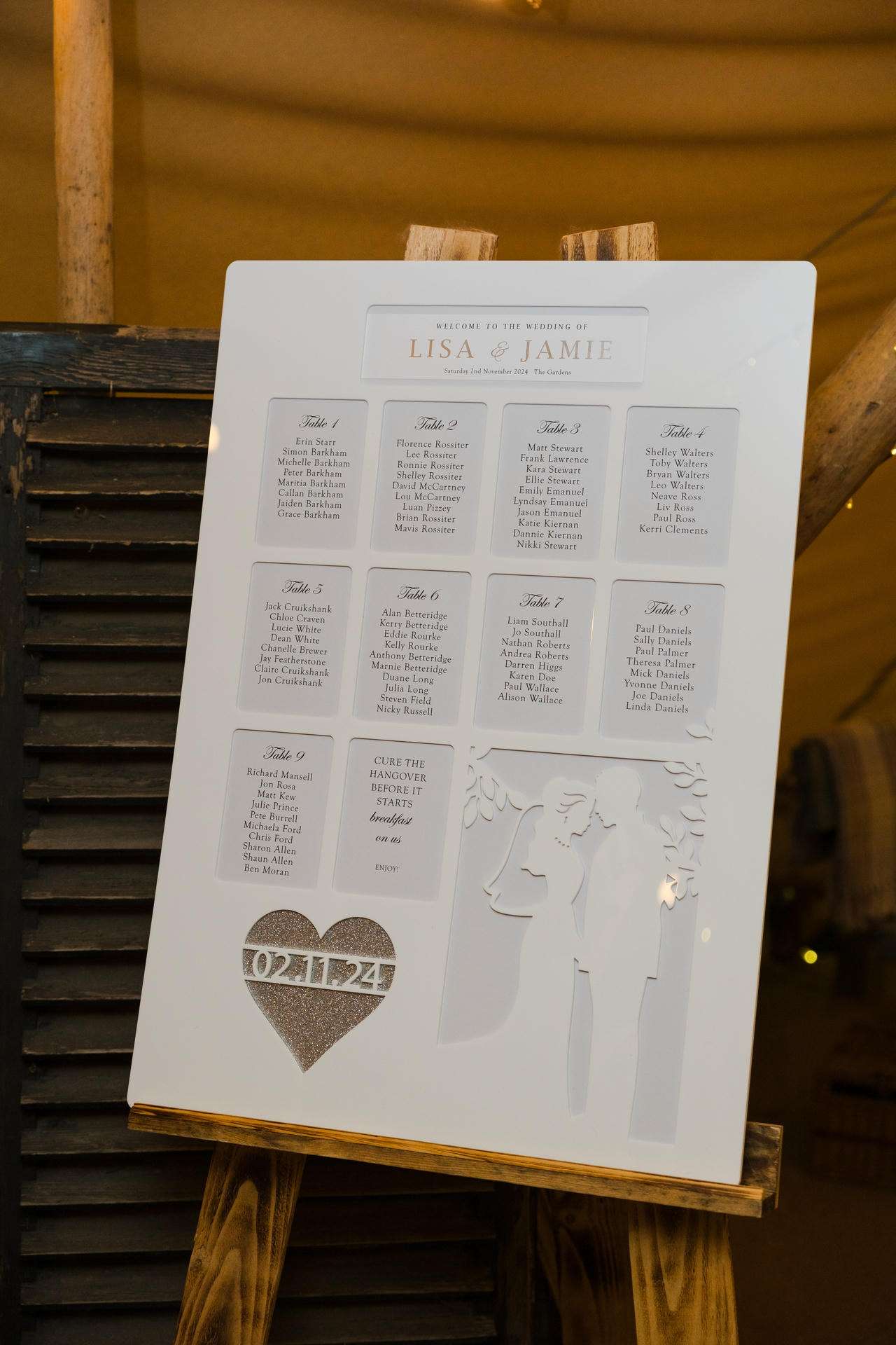 White wedding seating plan board on an easel showing Lisa & Jamie, table lists, a heart with the date, and a couple silhouette.