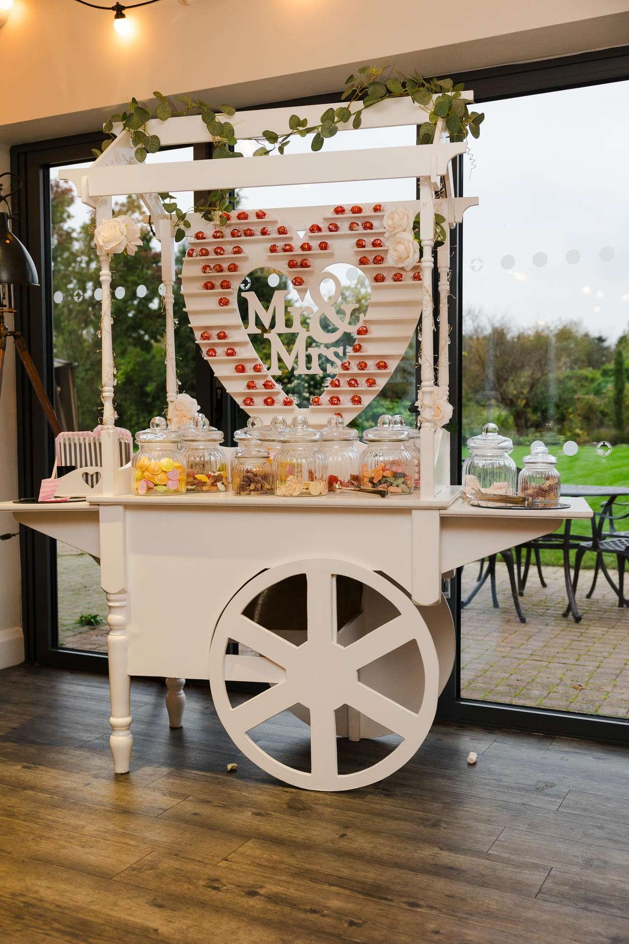 White vintage dessert cart with a heart-shaped backdrop reading Mr & Mrs, decorated with greenery and glass jars of candy.