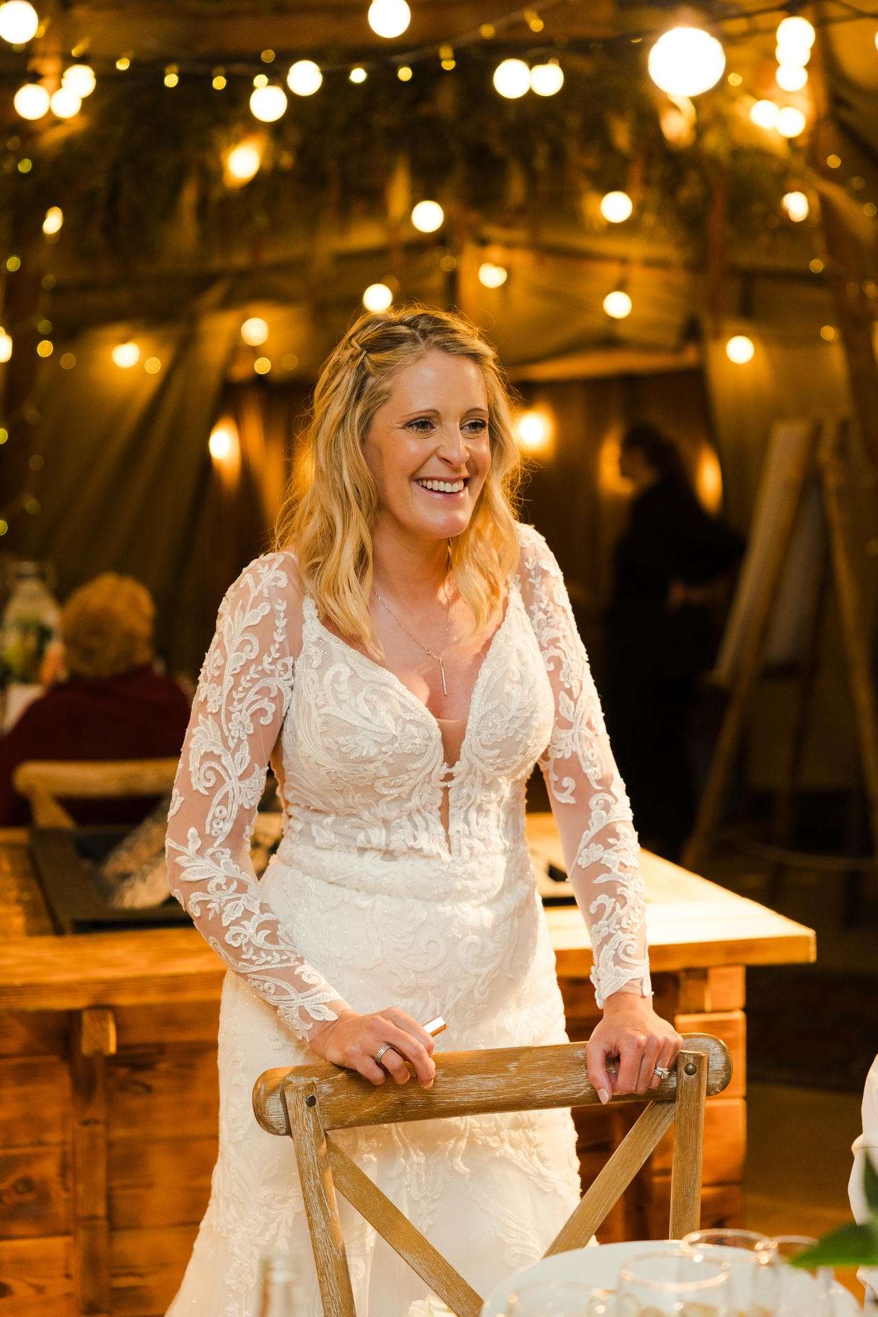Bride in a white lace wedding dress smiles as she stands beside a wooden chair under warm string lights.