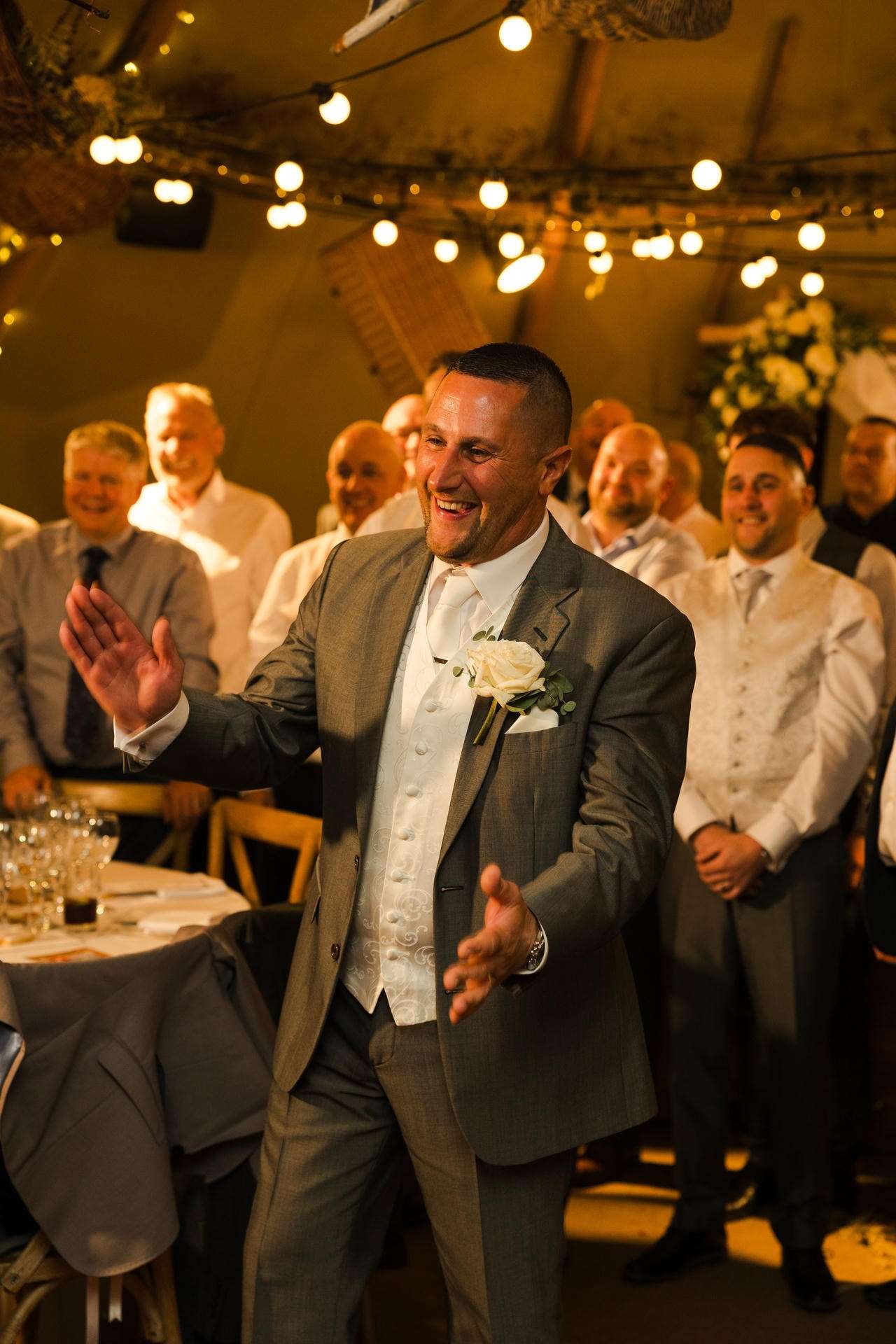 A man in a gray suit with a white boutonniere smiles and gestures while dancing at a wedding reception, with guests in the background.