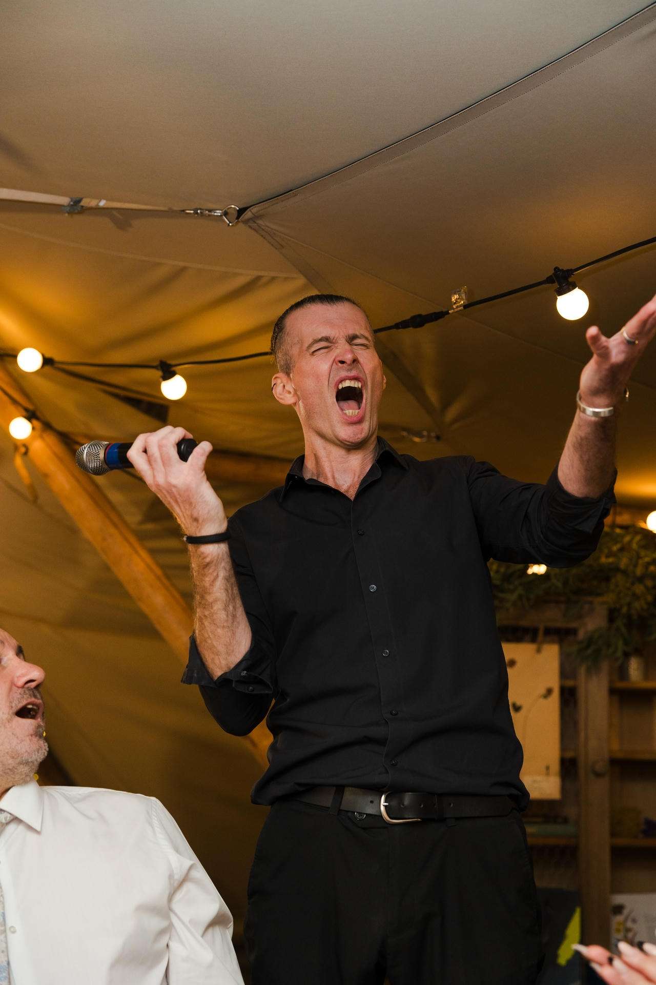Man in a black shirt passionately sings into a microphone under warm string lights inside a tent.