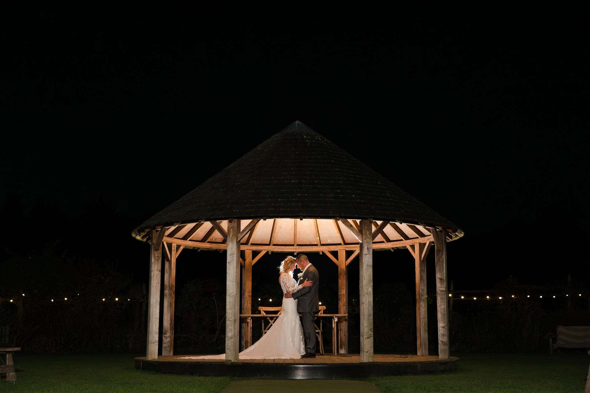 A bride and groom share a kiss under a warmly lit wooden gazebo at night.