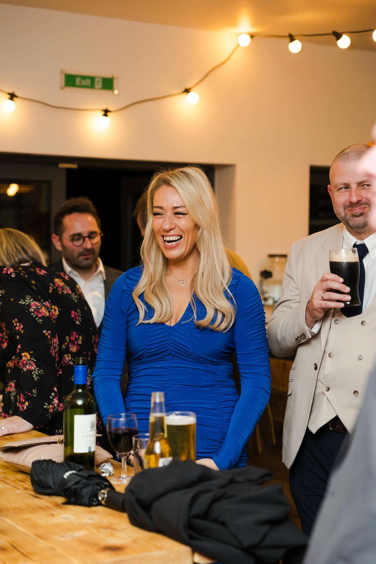 A smiling blonde woman in a blue dress stands by a wooden table with drinks at a lively party under string lights.