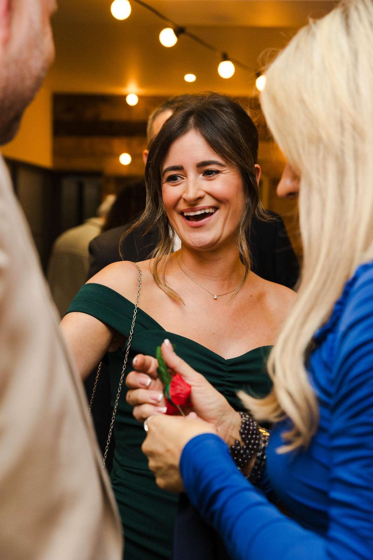 Smiling woman in an off-the-shoulder green dress chats with friends at a warmly lit social gathering.