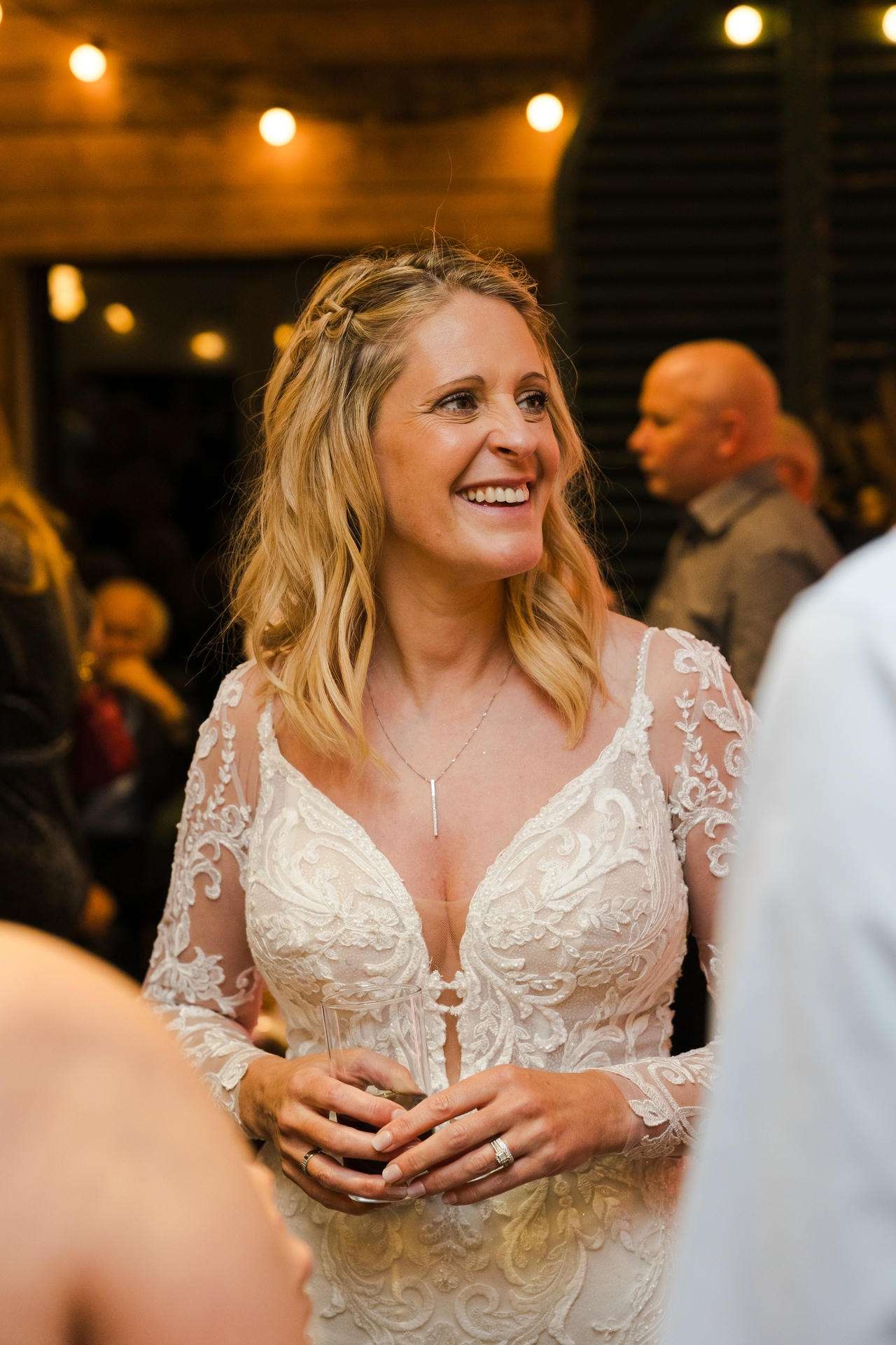 Smiling bride in a white lace wedding gown holds a glass at a reception.