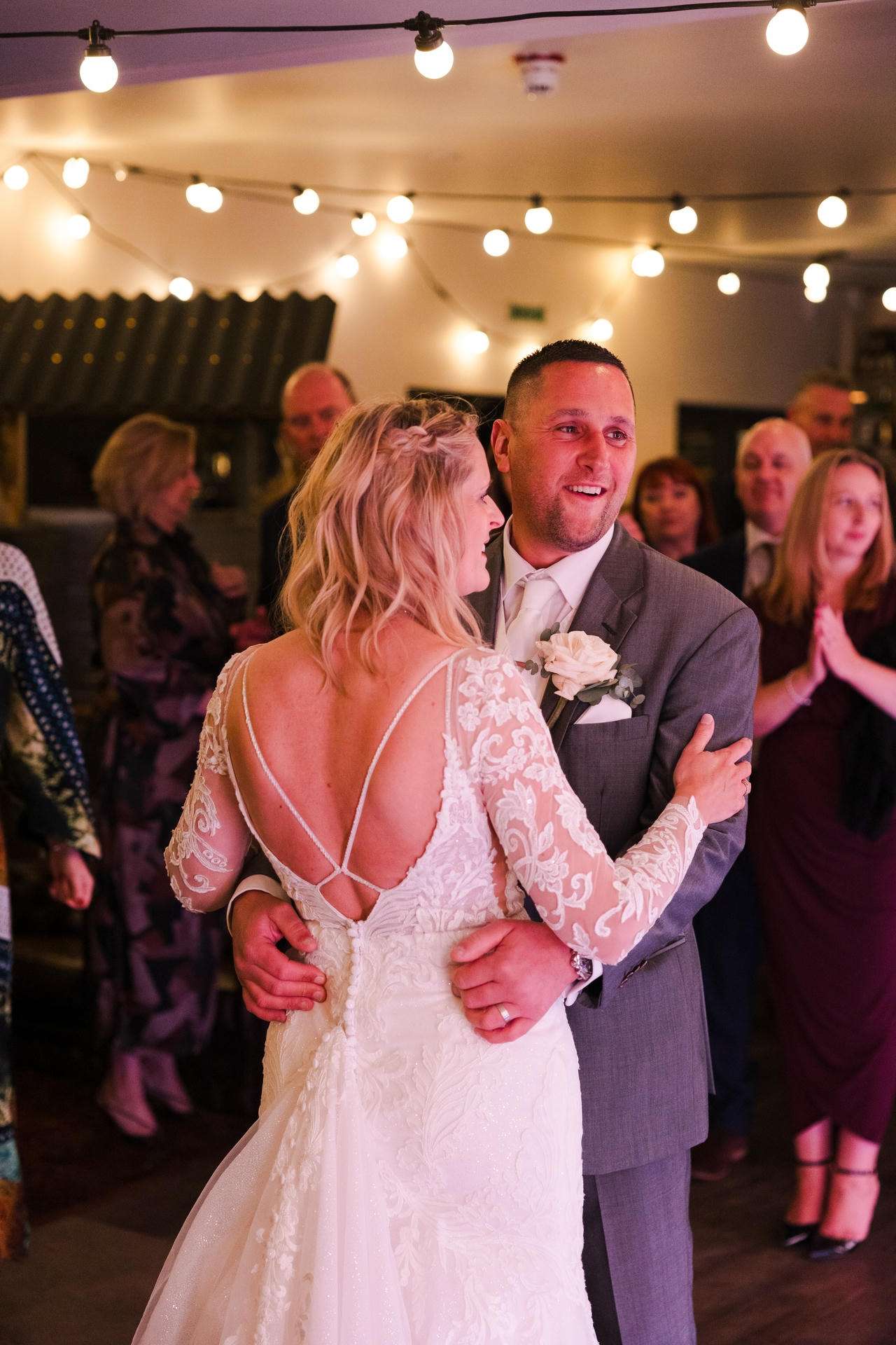 Bride in a lace wedding dress and groom in a gray suit dance together at their wedding reception under string lights.
