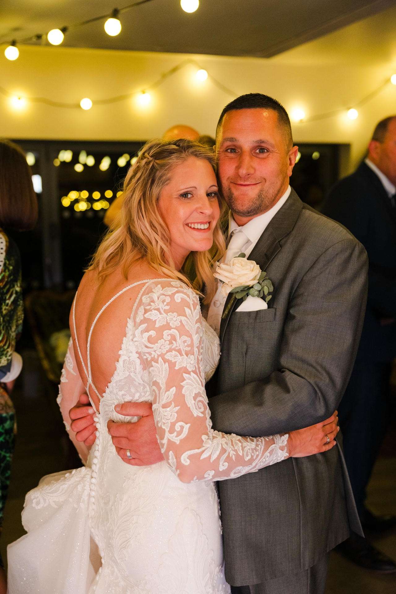 Bride in a long-sleeve lace wedding gown embraces her groom in a gray suit under warm string lights.
