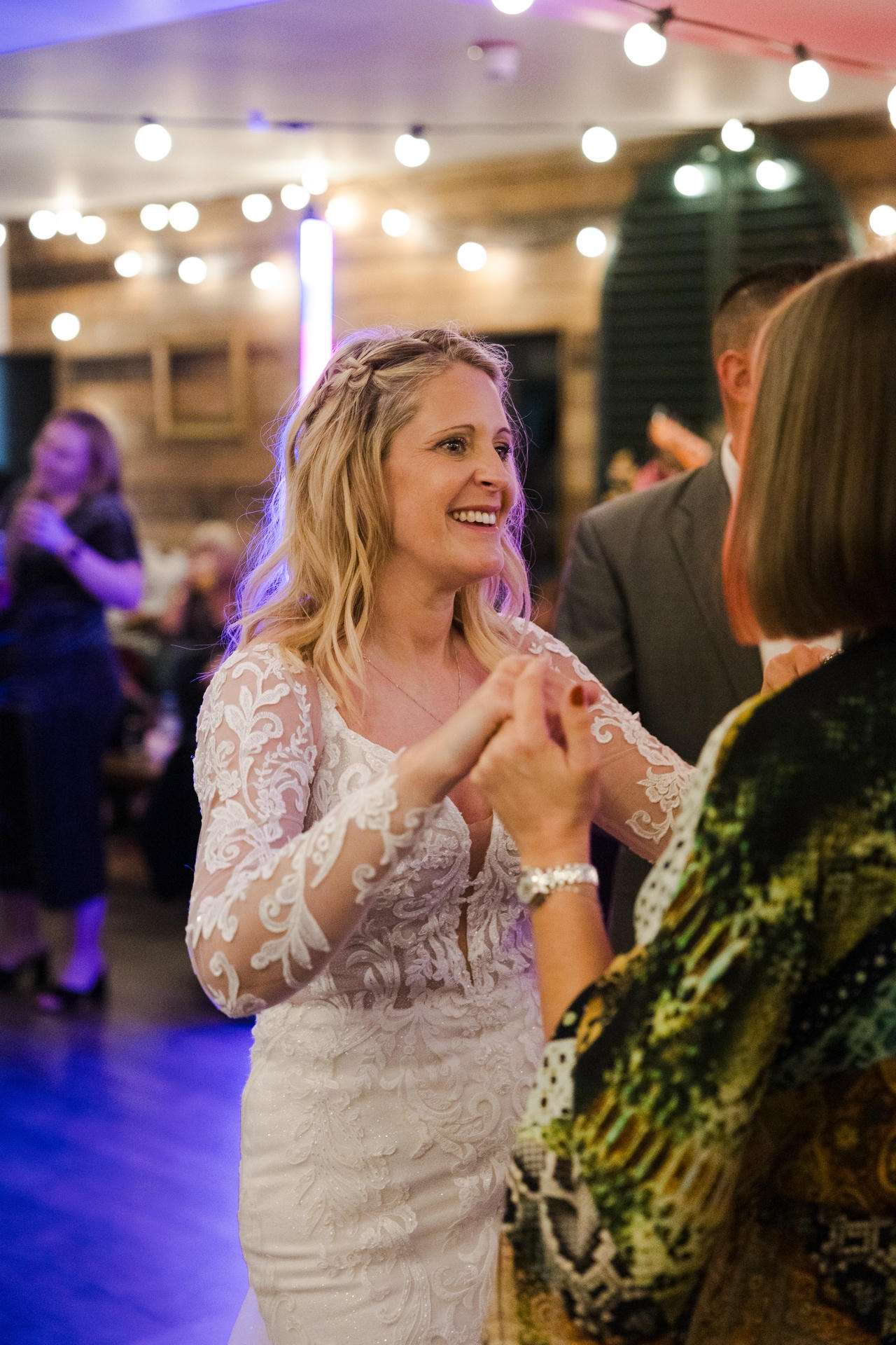 Smiling bride in a lace wedding gown dances with a guest at a warmly lit wedding reception.