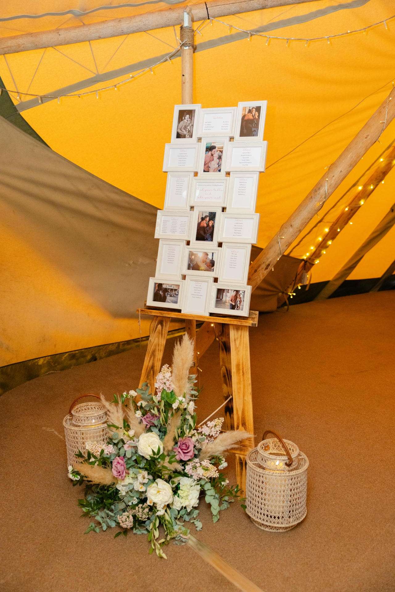 A rustic wooden stand displays a tower of white-framed photos inside a yellow tent, with a floral arrangement and wicker lanterns at its base.