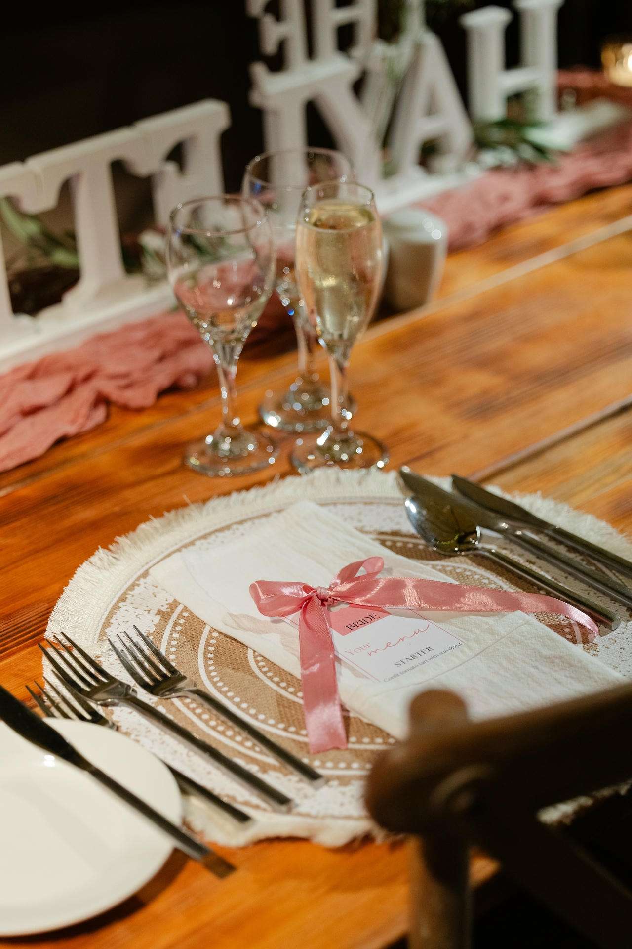 Wedding reception place setting with a pink ribbon-tied napkin on a decorative charger, plus silverware and champagne flutes.