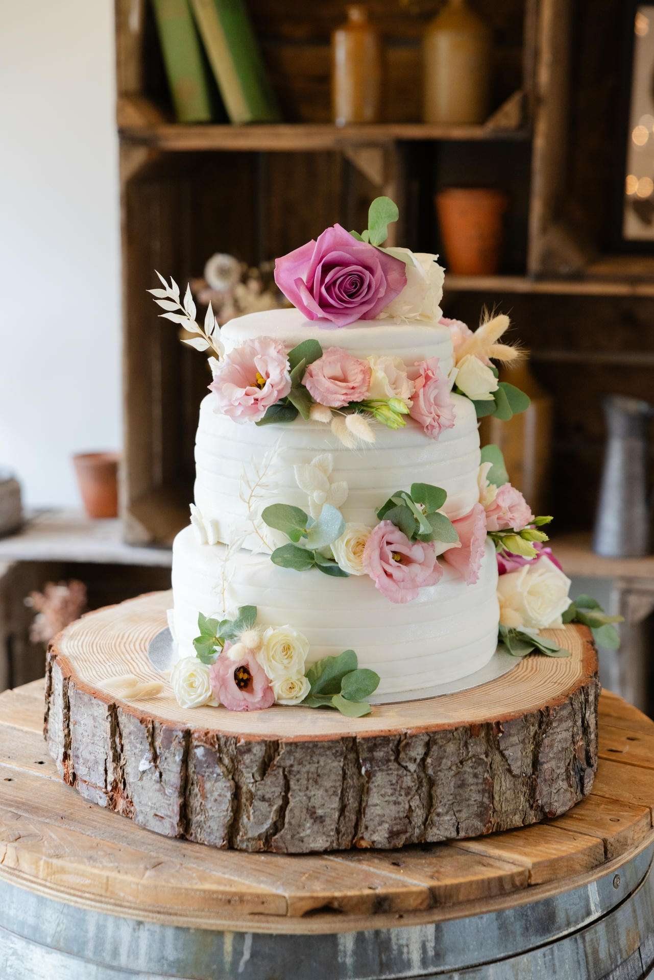 Three-tier white cake decorated with pink and white flowers and greenery, topped with a purple rose, on a rustic tree-stump cake stand.