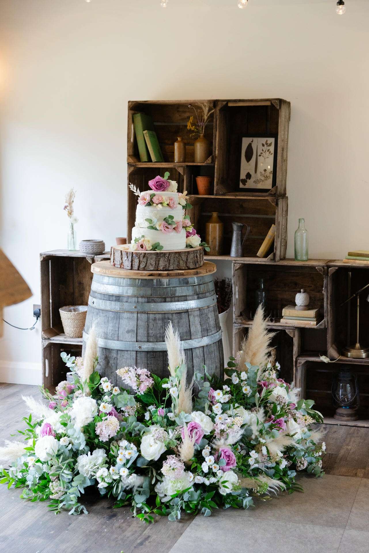 A white, tiered wedding cake adorned with pink flowers sits on a wooden barrel, surrounded by a lush floral arrangement in a rustic room.