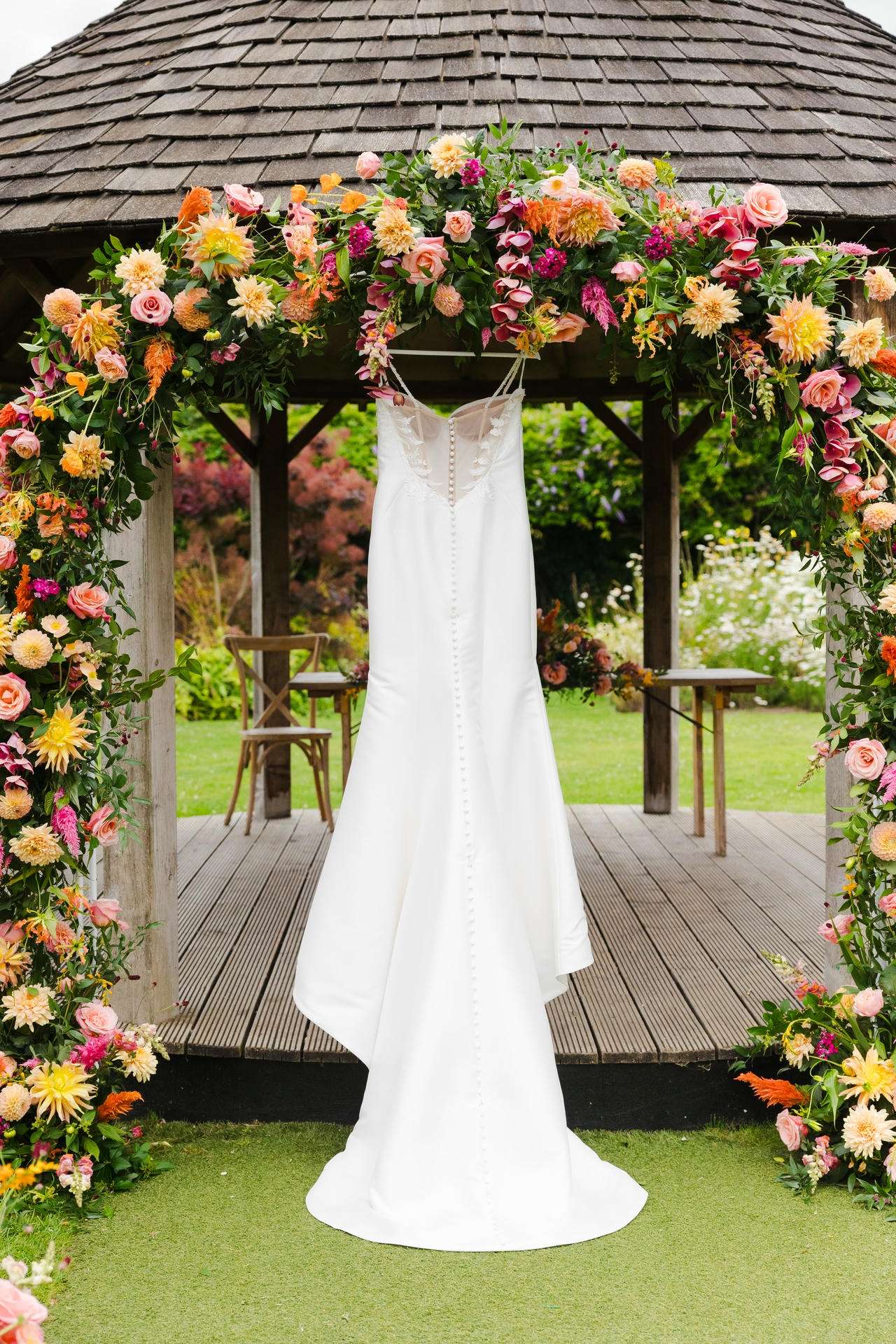 White wedding dress hanging from a colorful flower arch under a wooden gazebo.