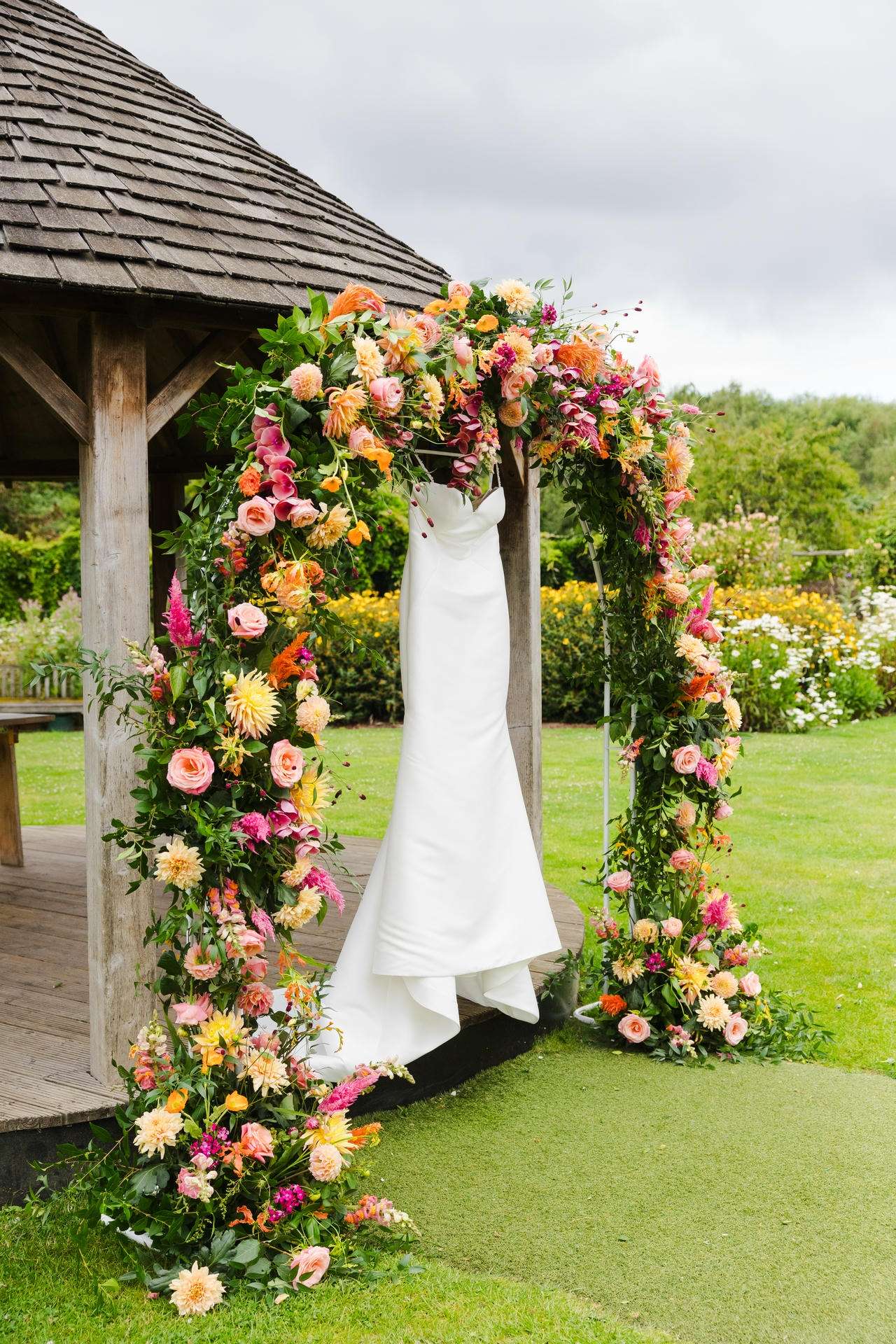 A white wedding dress hangs on a colorful flower arch beside a wooden garden gazebo.