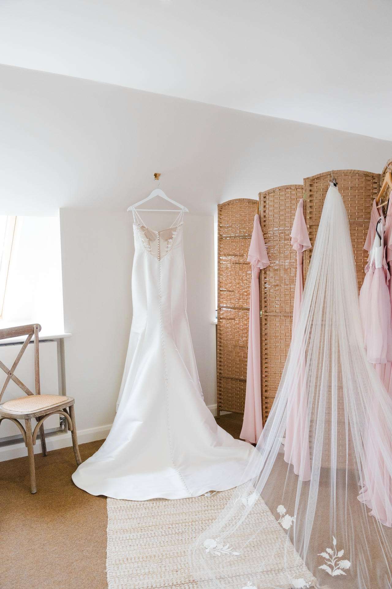 White wedding gown with a long train hangs on a hanger in a bright bridal fitting room, with a sheer veil trailing on the floor and pink dresses along a wicker divider.