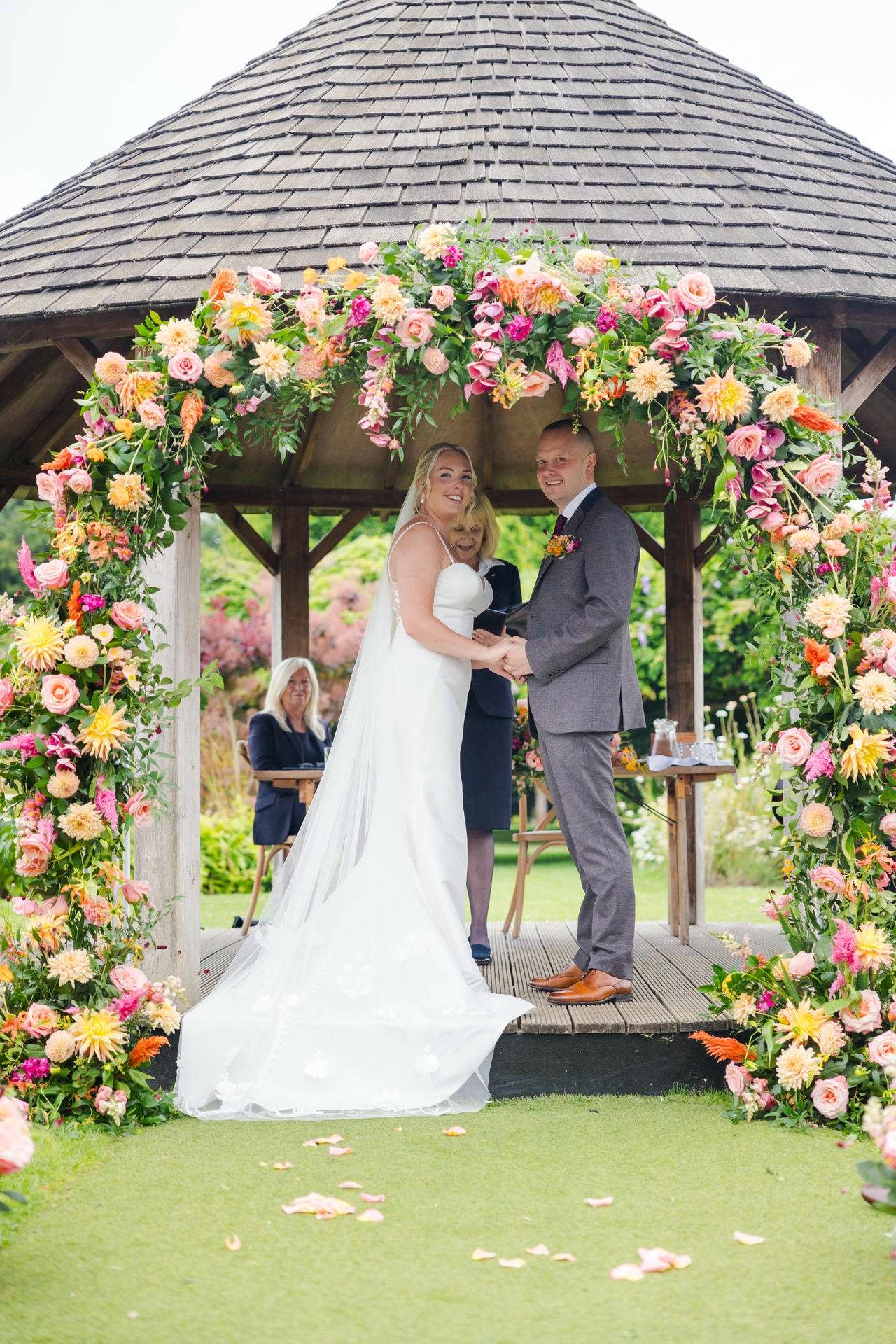 Bride and groom stand together under a vibrant flower arch in a wooden gazebo during their wedding ceremony.