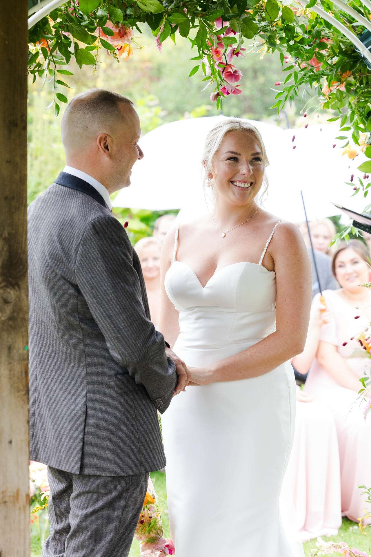 Bride and groom smile at each other while holding hands beneath a floral arch.