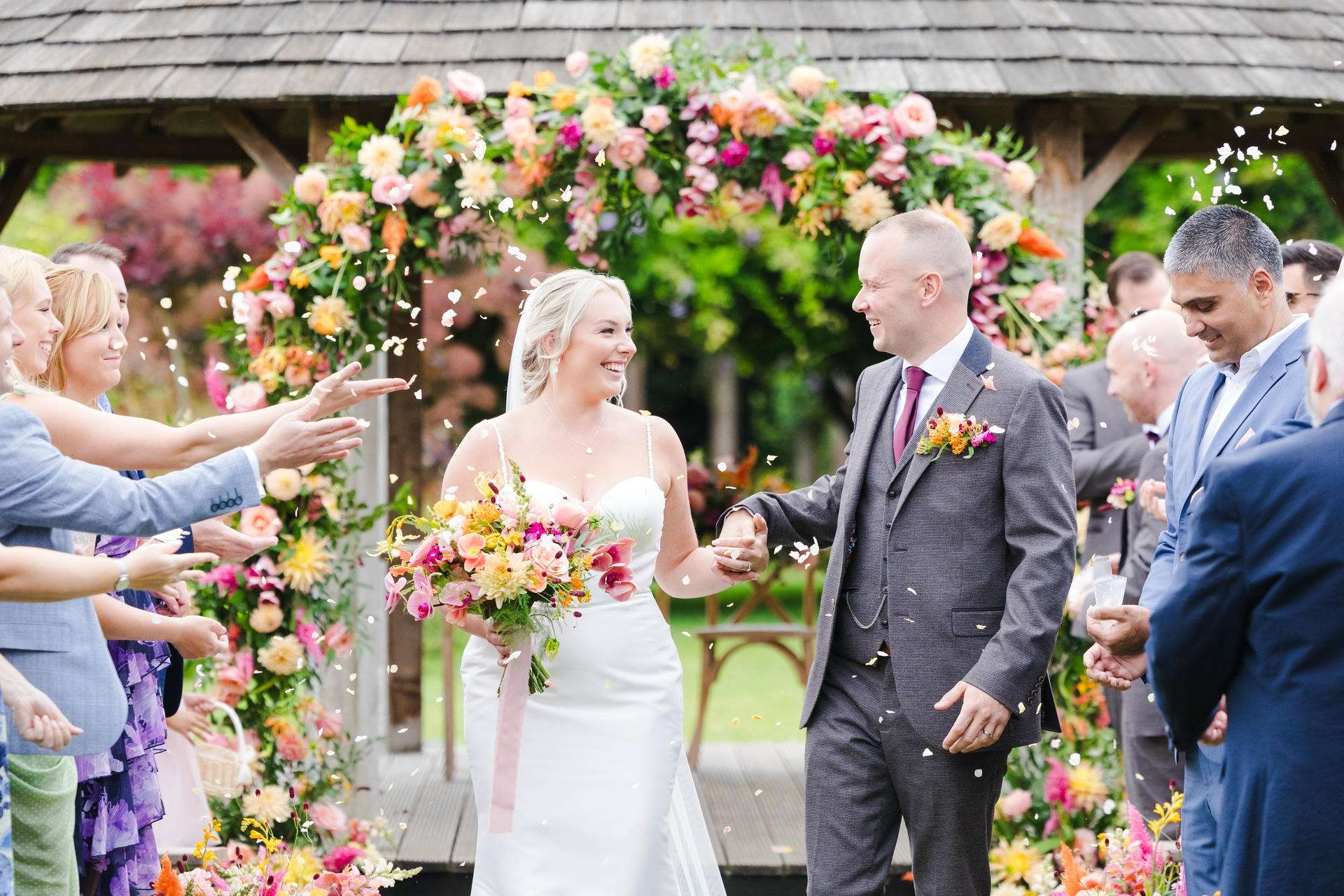 Bride and groom smile as guests shower them with flower petals under a colorful floral arch.