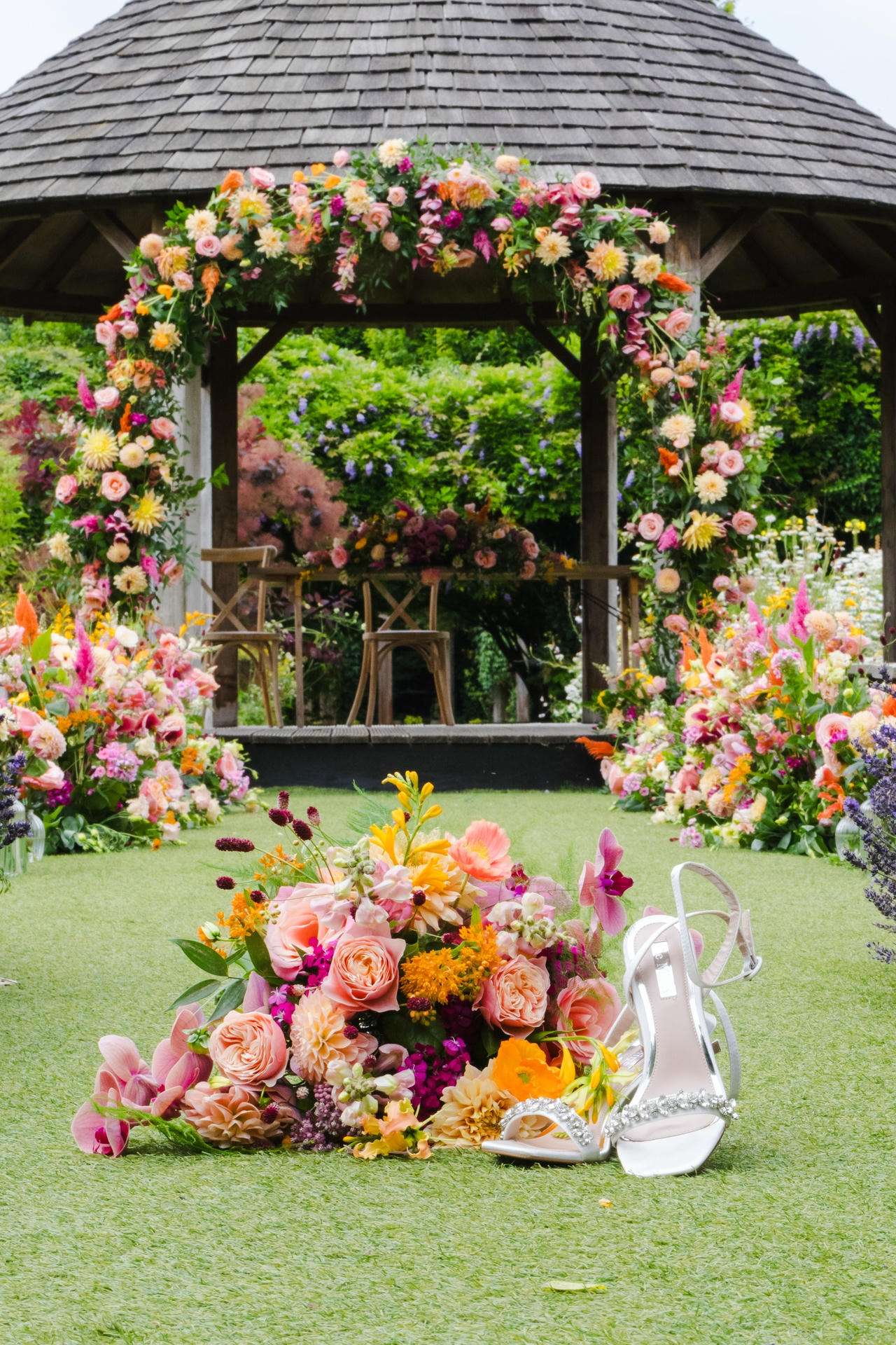 A vibrant floral arch frames a wooden gazebo in a garden, with colorful bouquets and a pair of white high-heeled shoes on the grass.