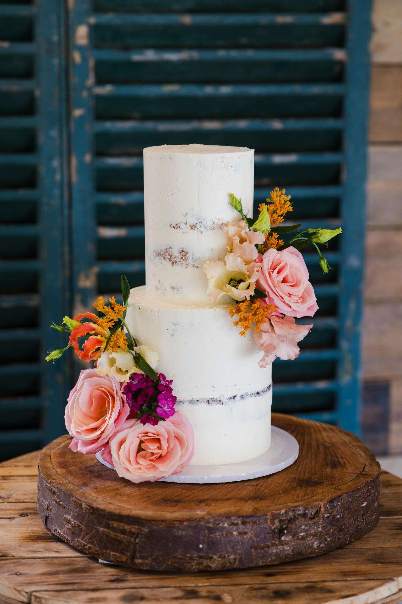 Three-tier naked white wedding cake decorated with pink roses and orange flowers, displayed on a round wooden cake stand on a rustic wooden table.