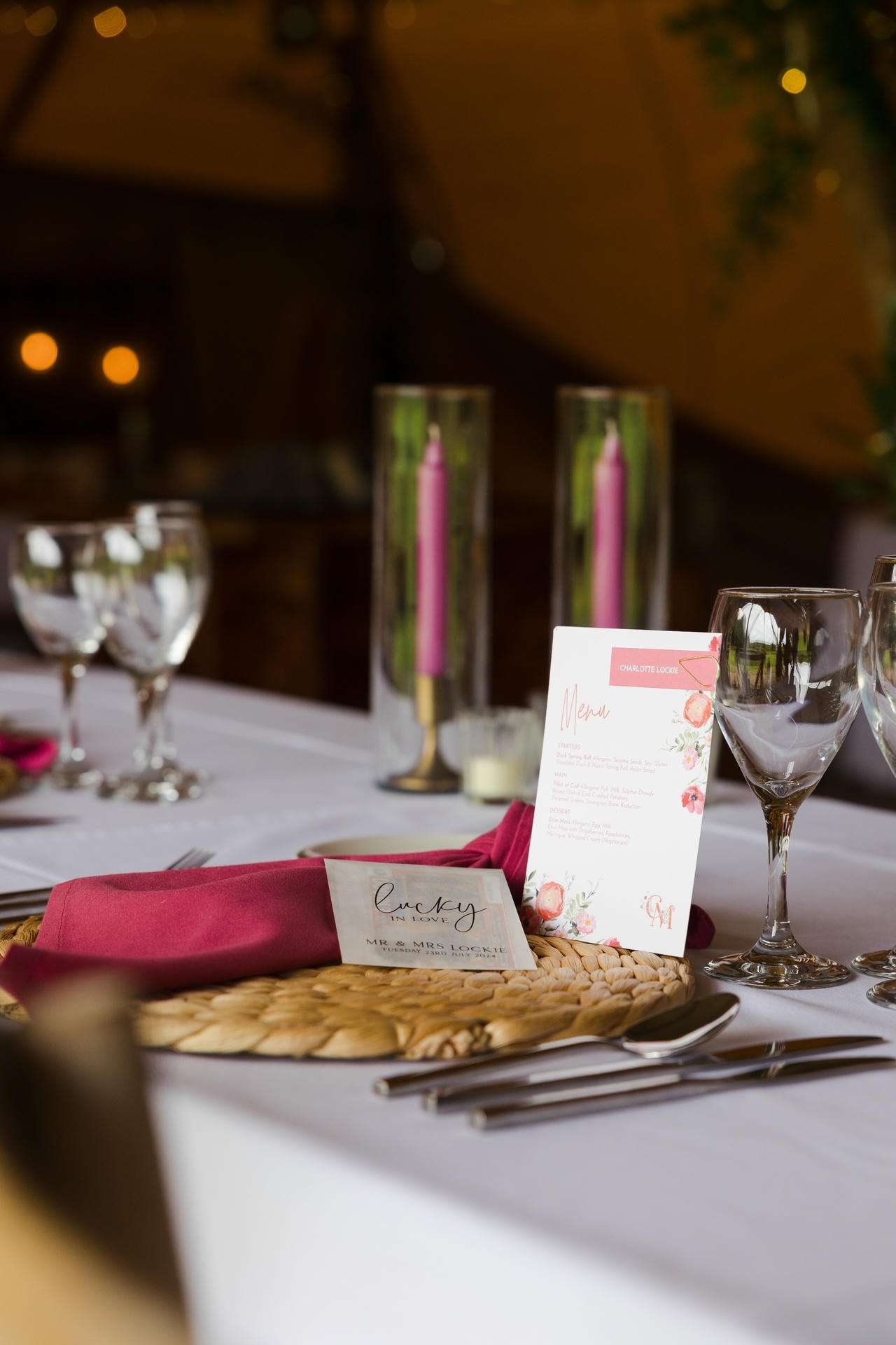 Elegant table setting featuring a woven placemat, pink napkin, name card, menu, wine glasses, and tall pink candles in glass holders.