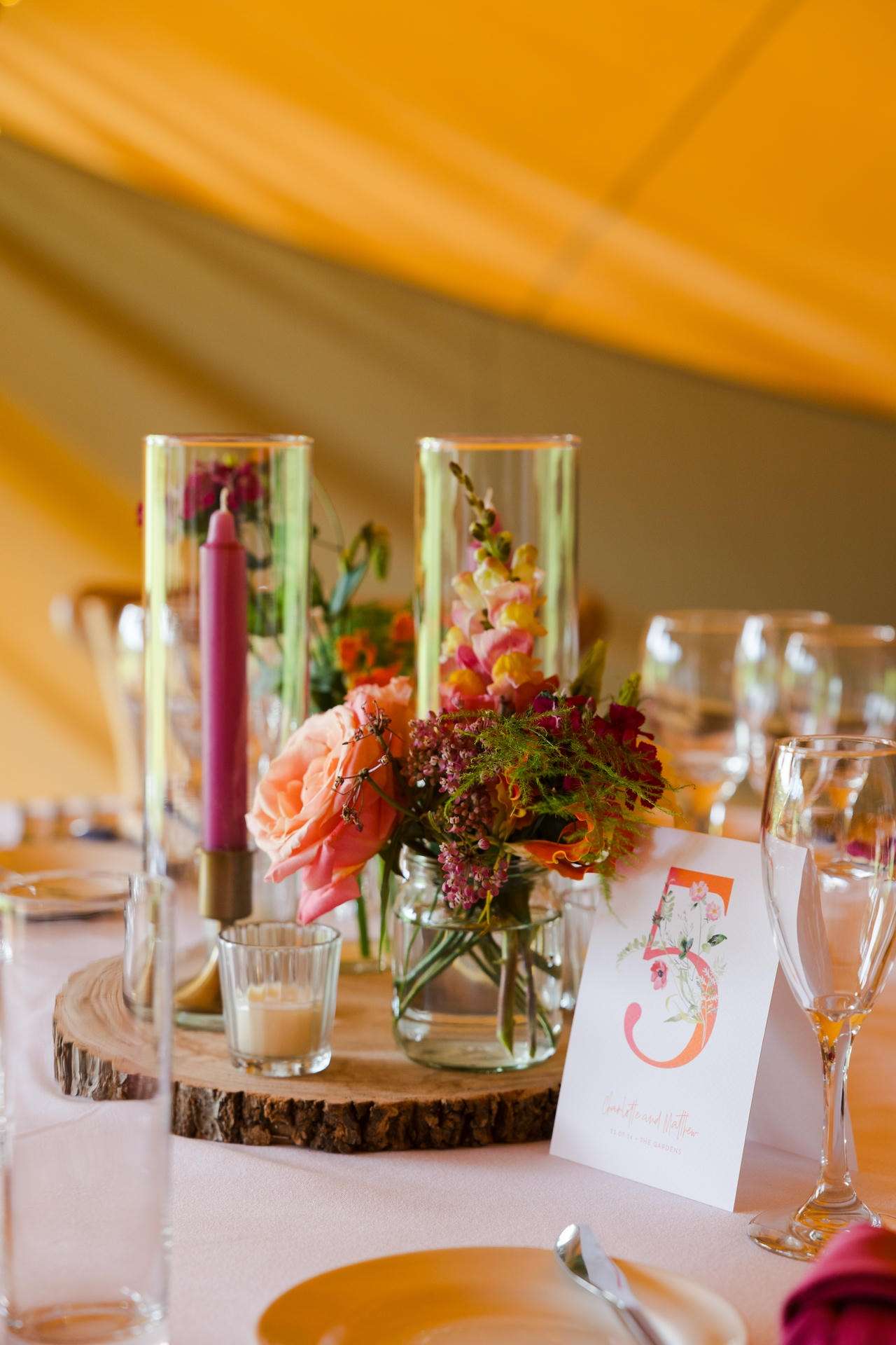 Table laid for a celebration with a flower-filled centerpiece in glass jars on a wooden slab, a tall purple candle, and a card marked with the number 5.
