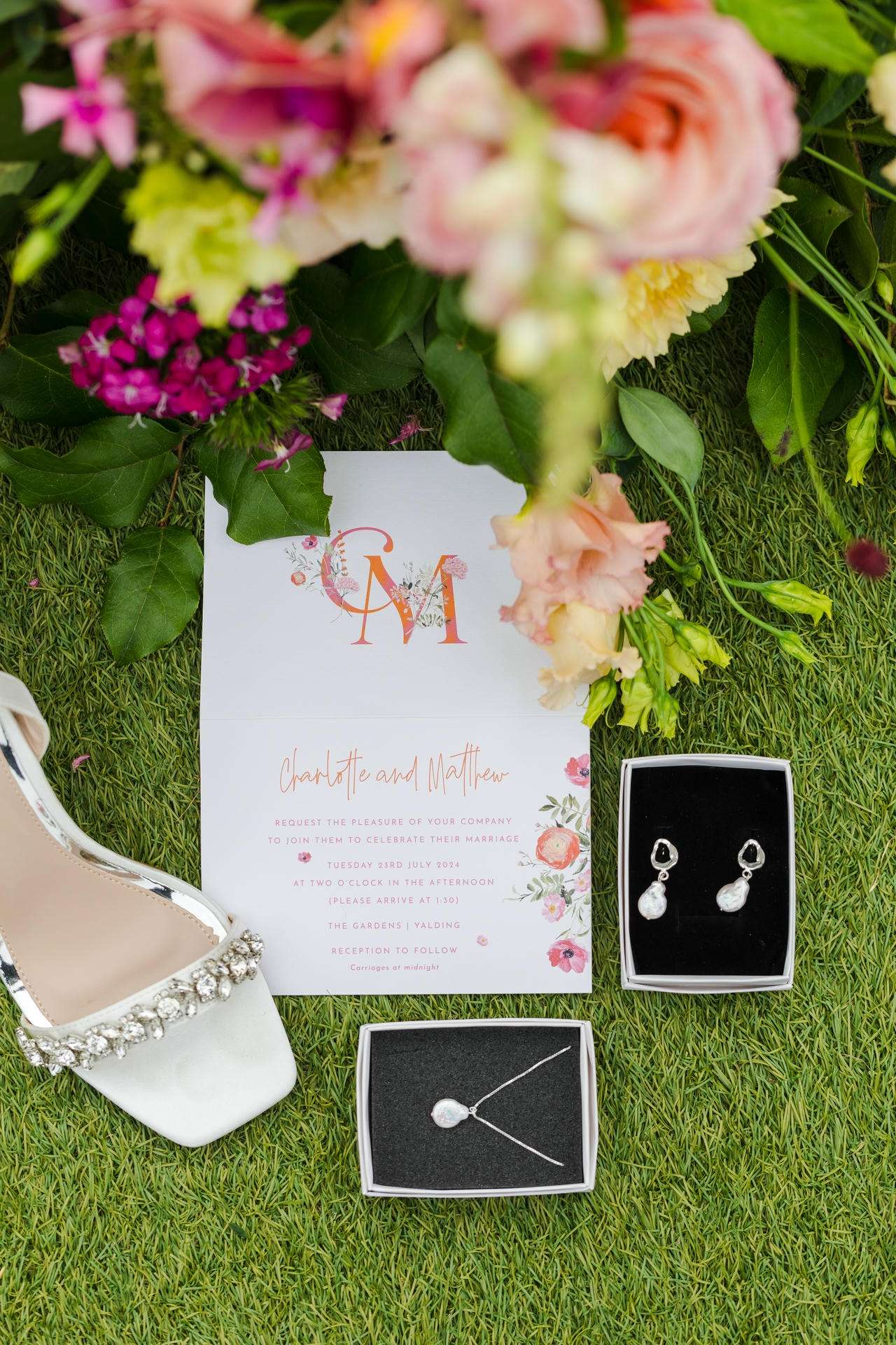Wedding invitation on green grass framed by a bouquet, with a white jeweled shoe and boxes holding a necklace and earrings.