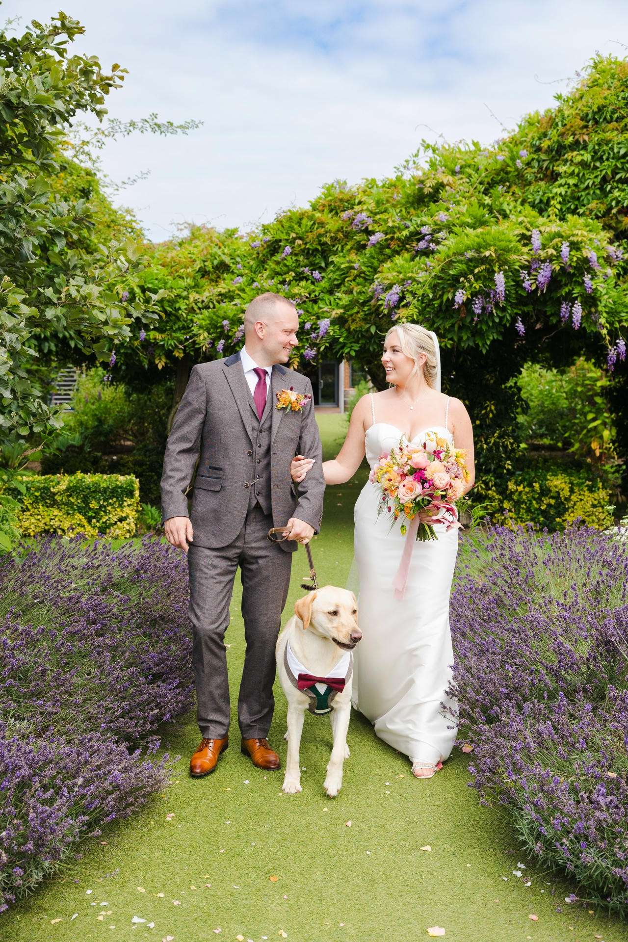 Bride and groom in wedding attire walk with their dog along a garden path lined with purple flowers at The Gardens Wedding venue