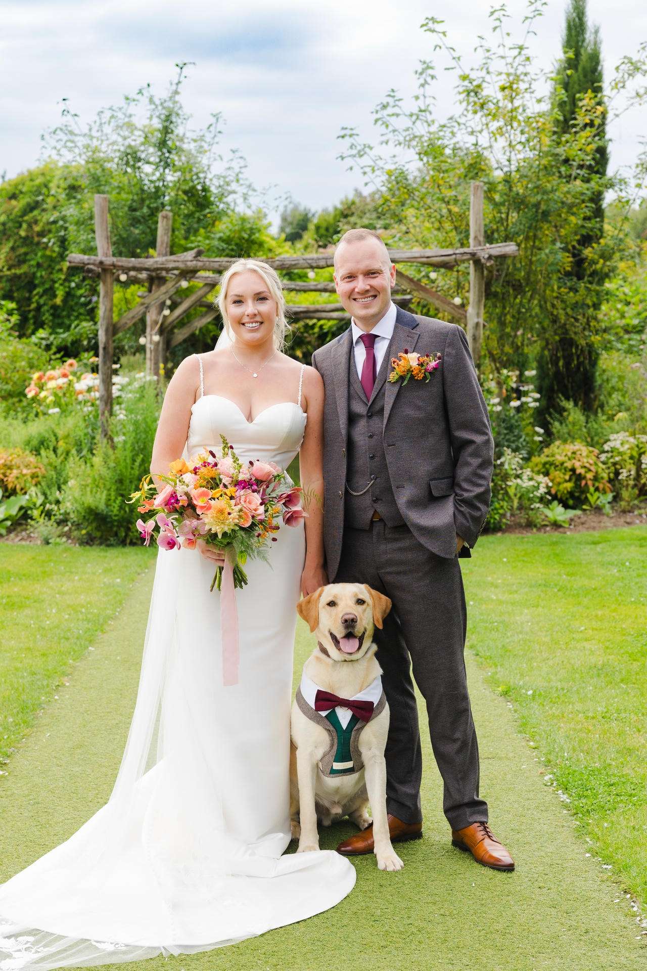 Bride in a white wedding gown and groom in a gray suit pose with a dog wearing a bow tie in a garden wedding at The Gardens Wedding venue