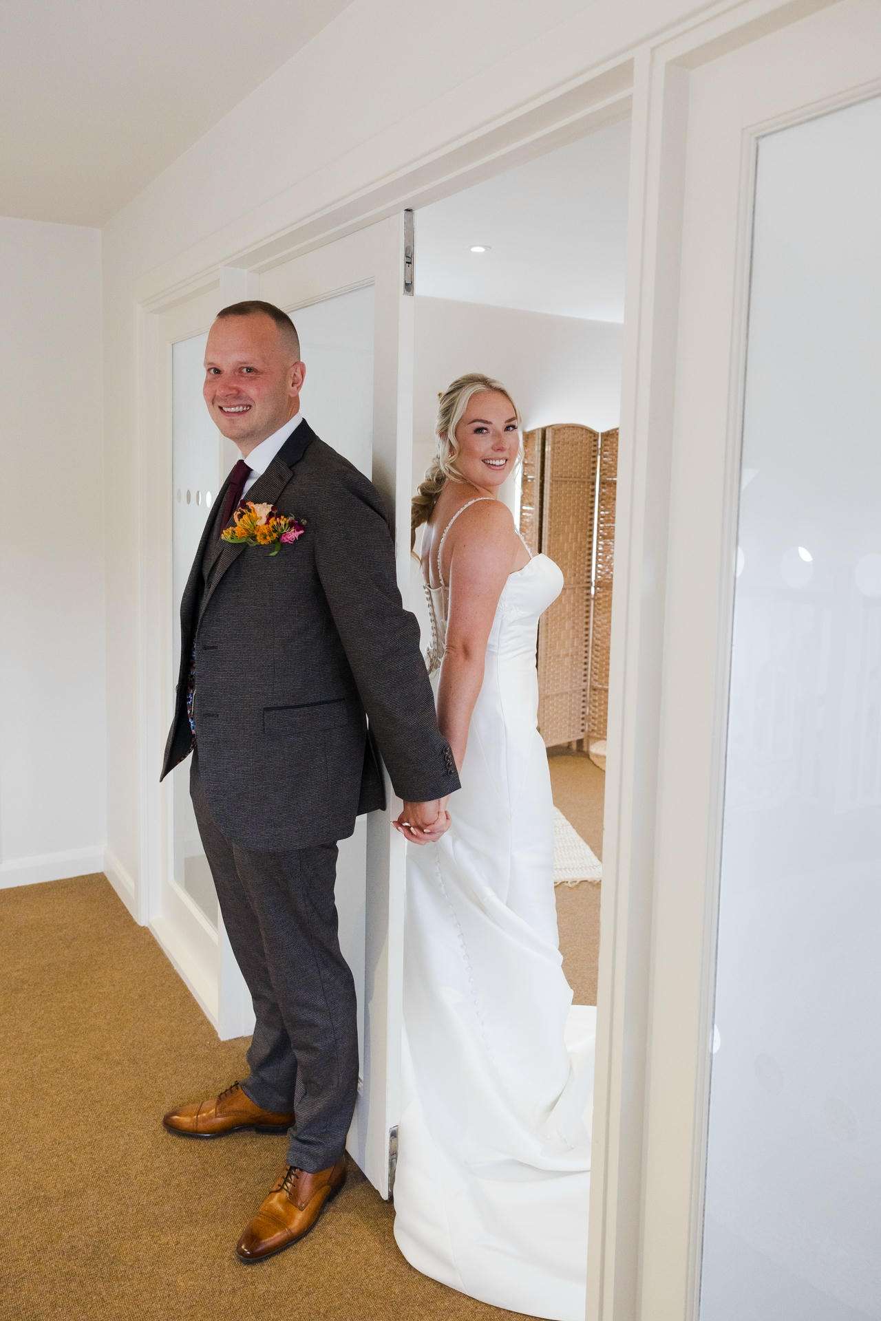 Smiling bride in a white wedding gown and groom in a dark gray suit stand back-to-back, holding hands in a doorway.