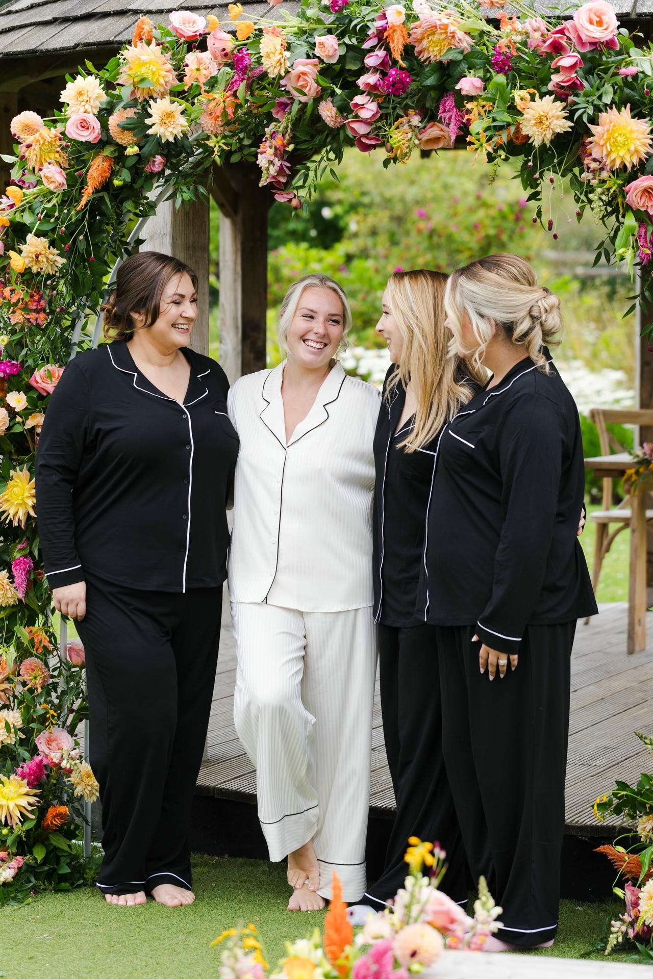 Four women in black and white pajamas stand barefoot under a vibrant flower arch outdoors, smiling at each other.