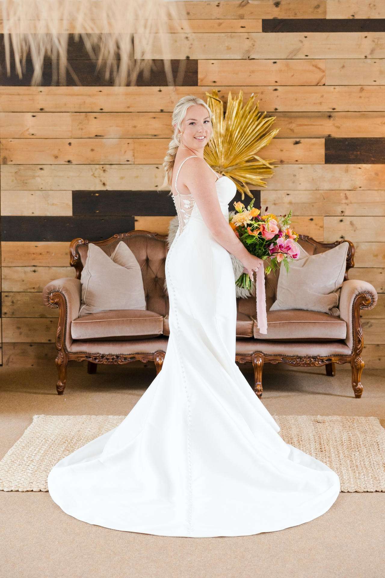 Bride in a white wedding gown with a long train stands with a colorful bouquet beside a vintage sofa against a wood-paneled wall.