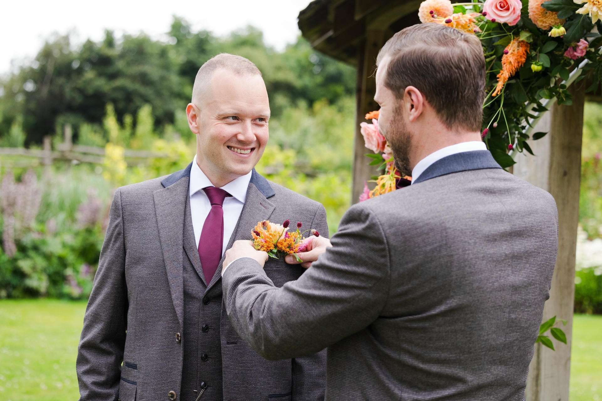 Two men in gray suits outdoors, one pinning a colorful boutonniere on the other's lapel and smiling.