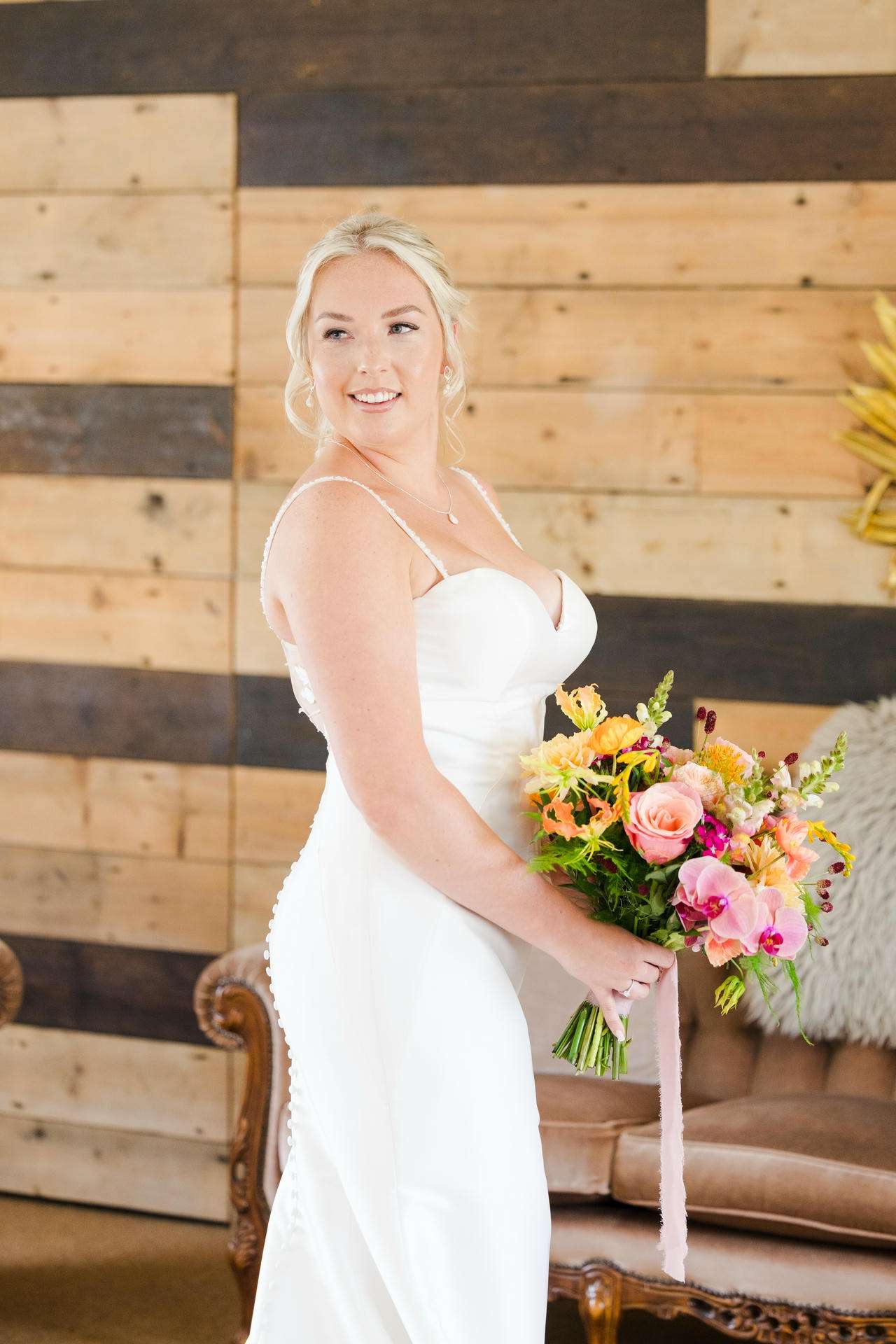 A smiling bride in a white wedding dress holds a bright bouquet of pink, orange, and yellow flowers indoors against a wood-paneled wall. at The Gardens Wedding venue