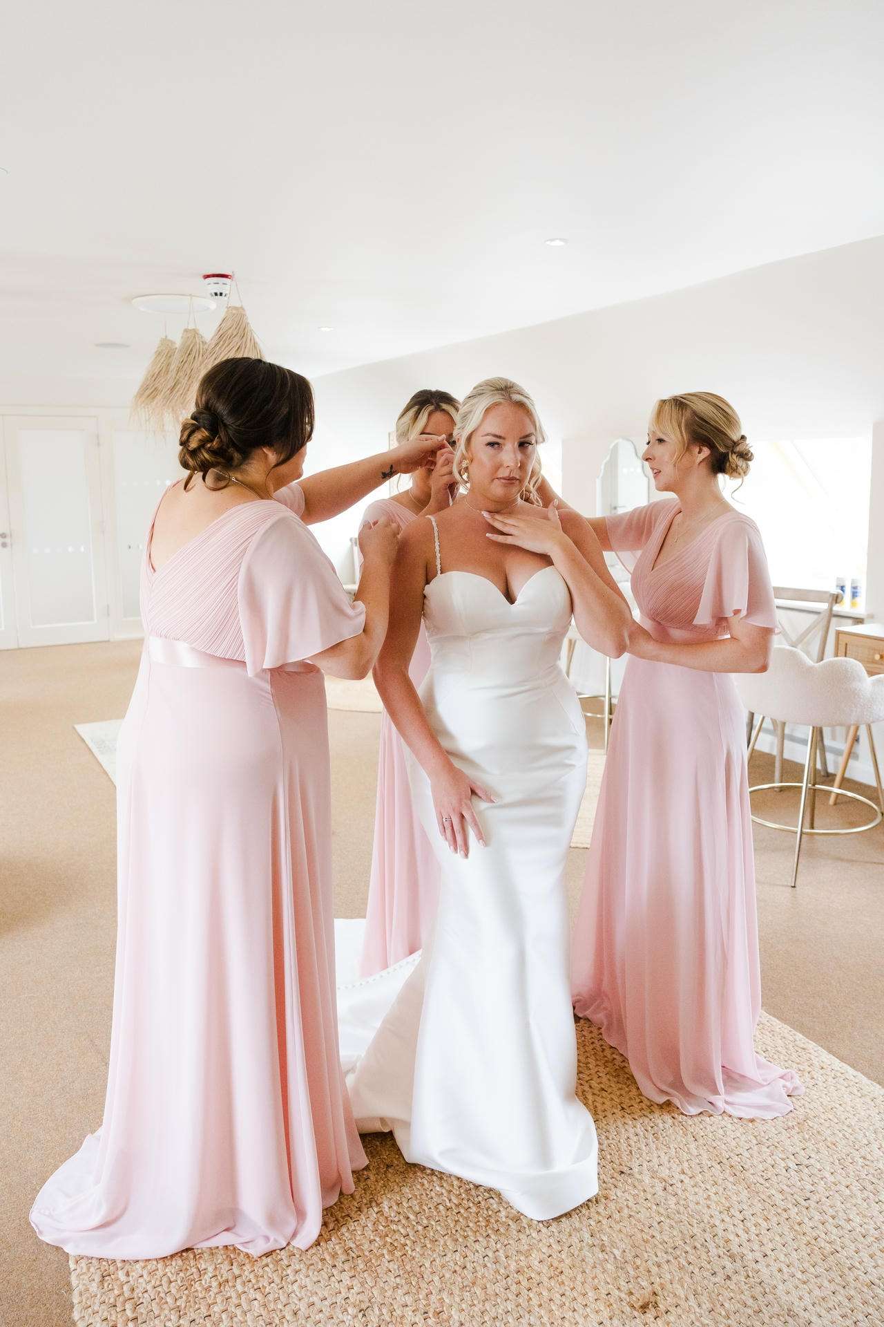 Bride in a white wedding gown is helped by three bridesmaids in pink dresses in a bright, airy room.