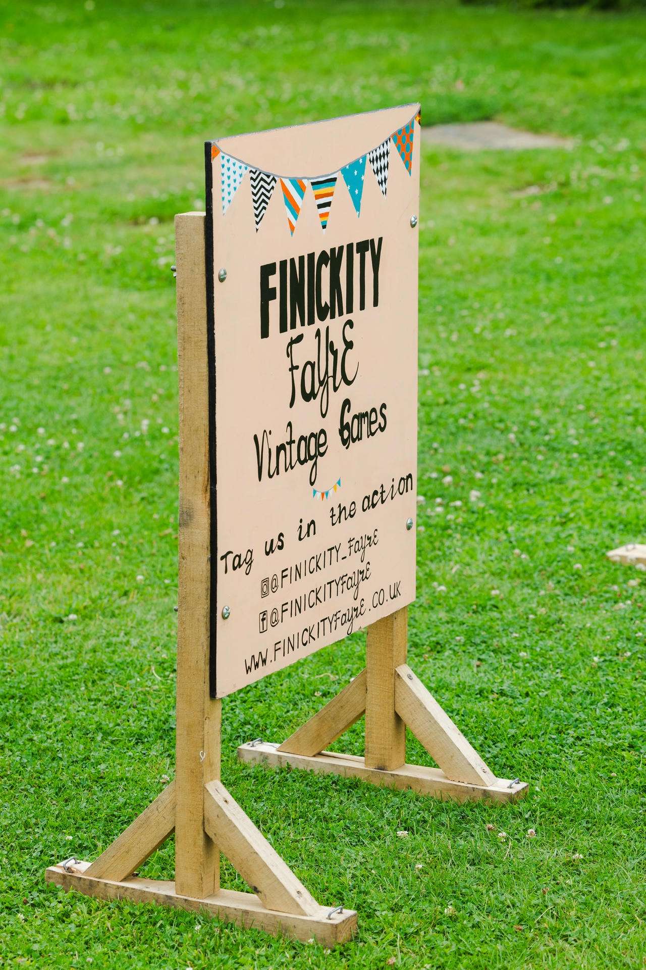 An A-frame wooden sign on a grassy field advertising Finickity Fayce Vintage Games with colorful pennant bunting.