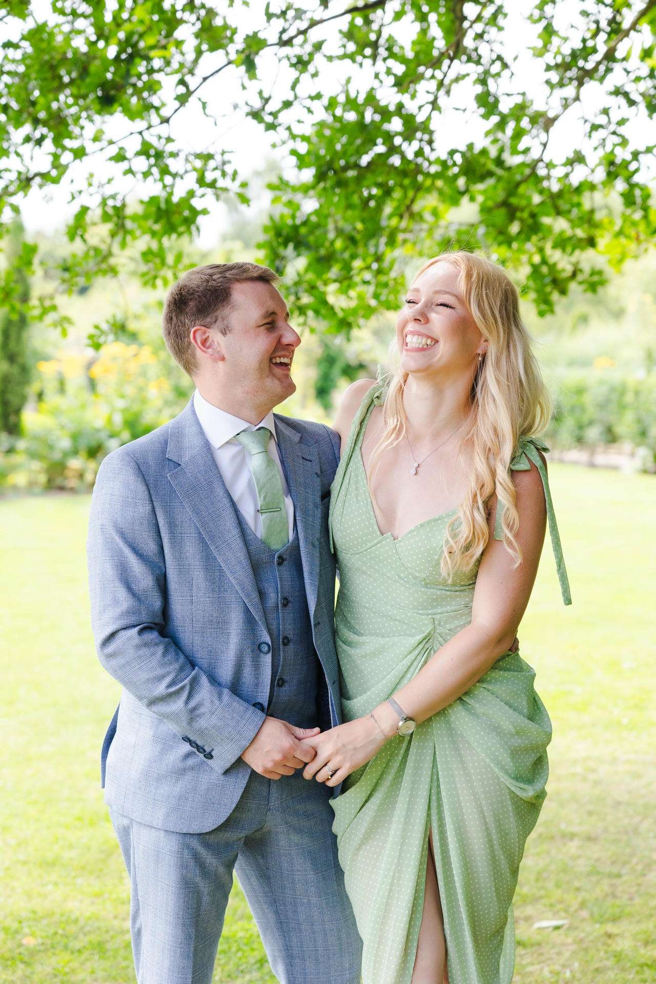 A smiling couple stands in a sunlit park, the man in a light blue suit and the woman in a mint green dress, smiling and holding hands.