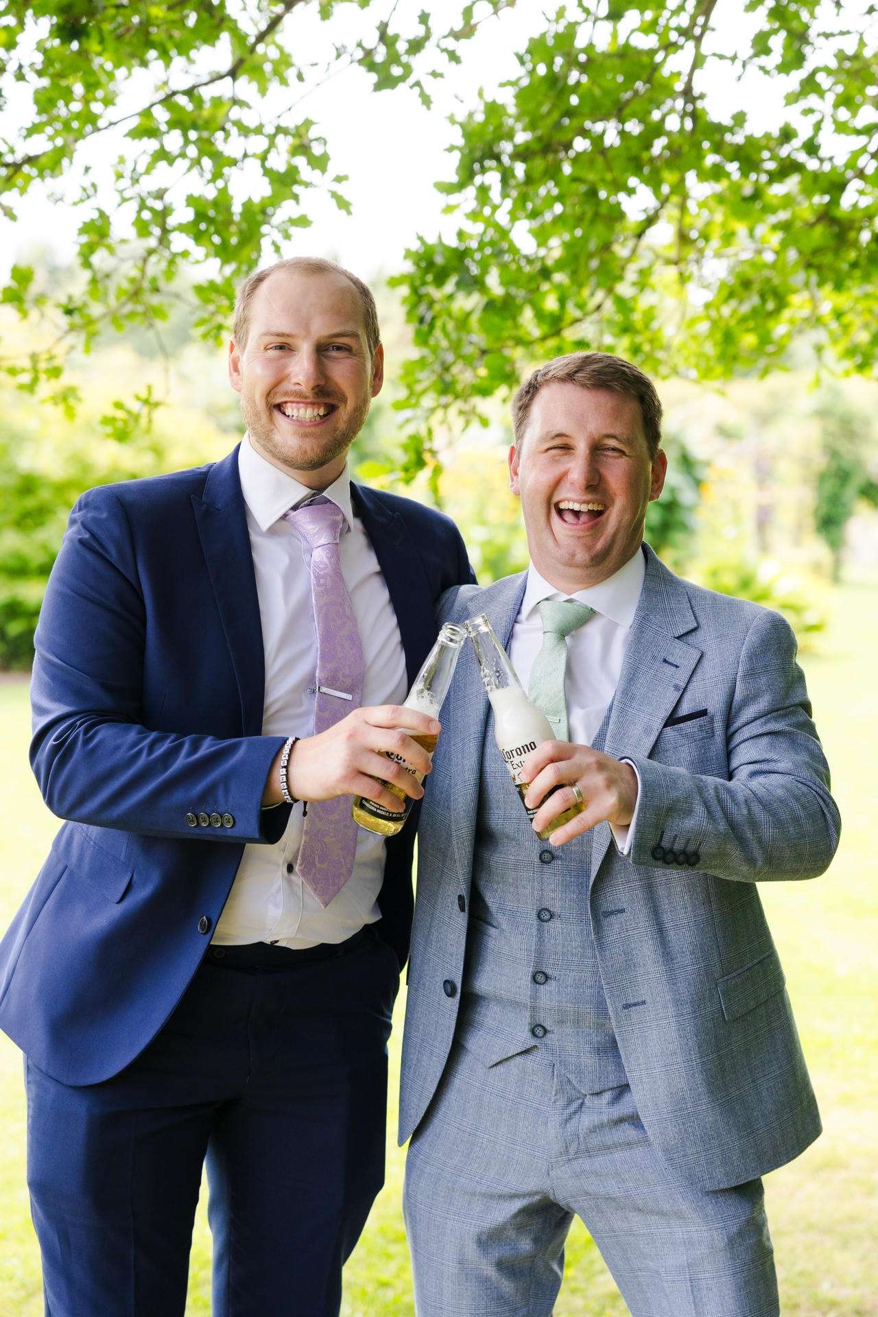 Two men in suits outdoors, smiling and toasting with beer bottles under leafy trees.