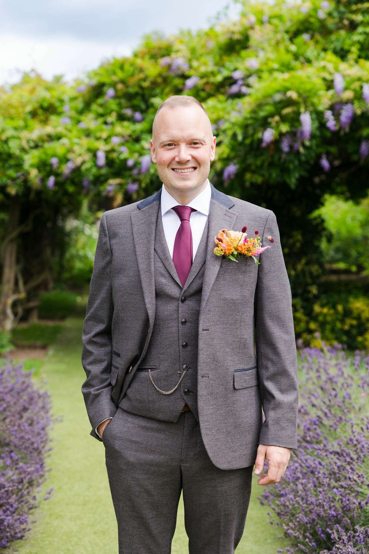 Smiling man in a gray checkered three-piece suit with a burgundy tie and floral boutonniere stands in a garden with purple flowering plants.