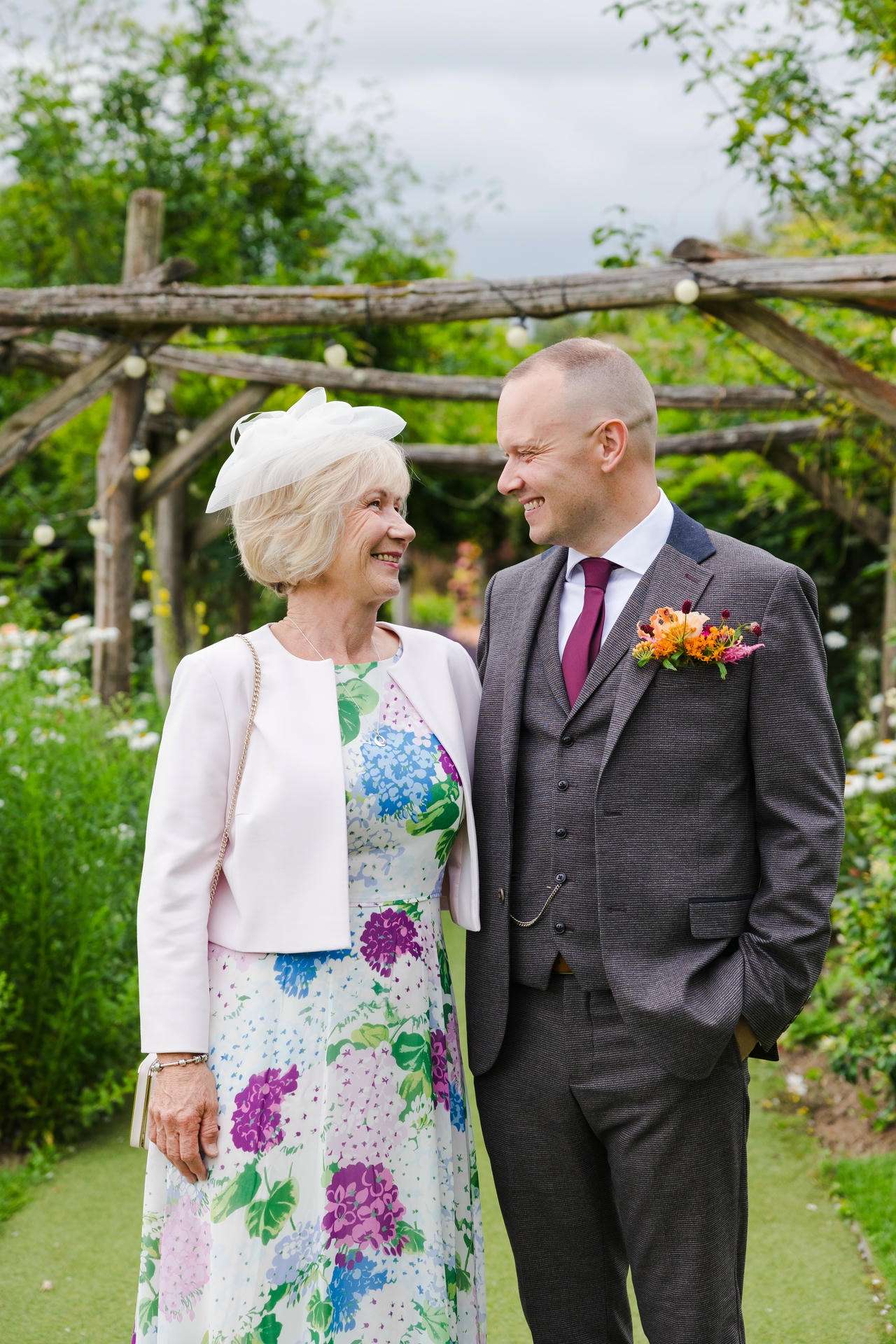 An older woman in a floral dress with a white fascinator and jacket stands beside a man in a gray suit with a boutonniere, smiling at each other in a garden.