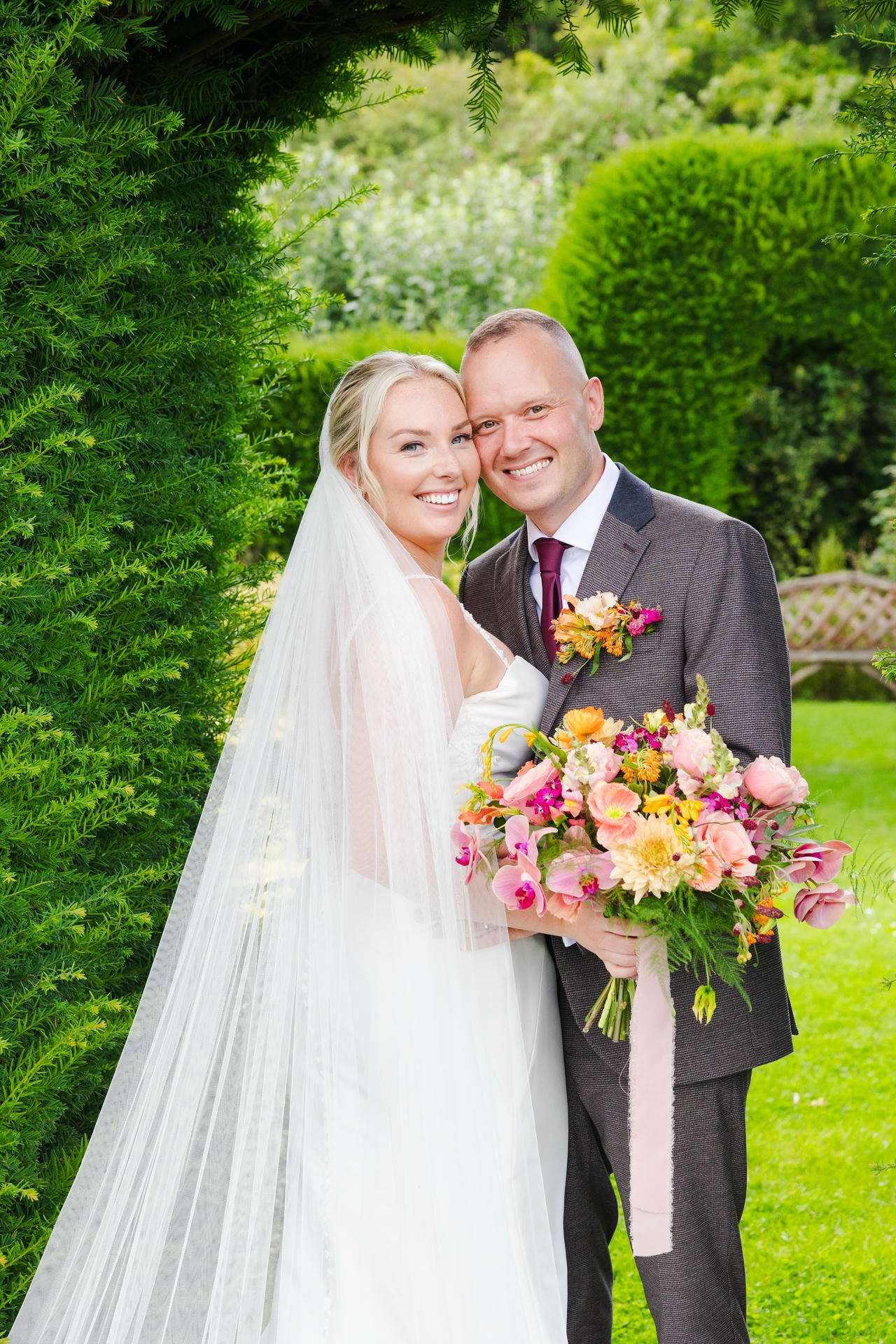 A smiling bride in a white wedding dress and veil stands beside her groom in a dark suit in a lush garden, holding a colorful bouquet at The Gardens Wedding venue